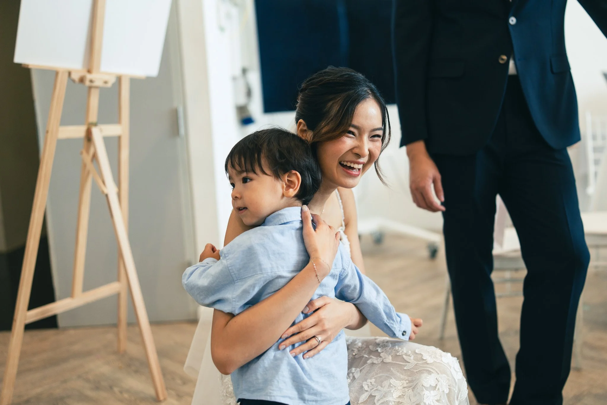A woman happily hugging a young boy in a light blue shirt in an indoor setting, with a person in a dark suit standing nearby.
