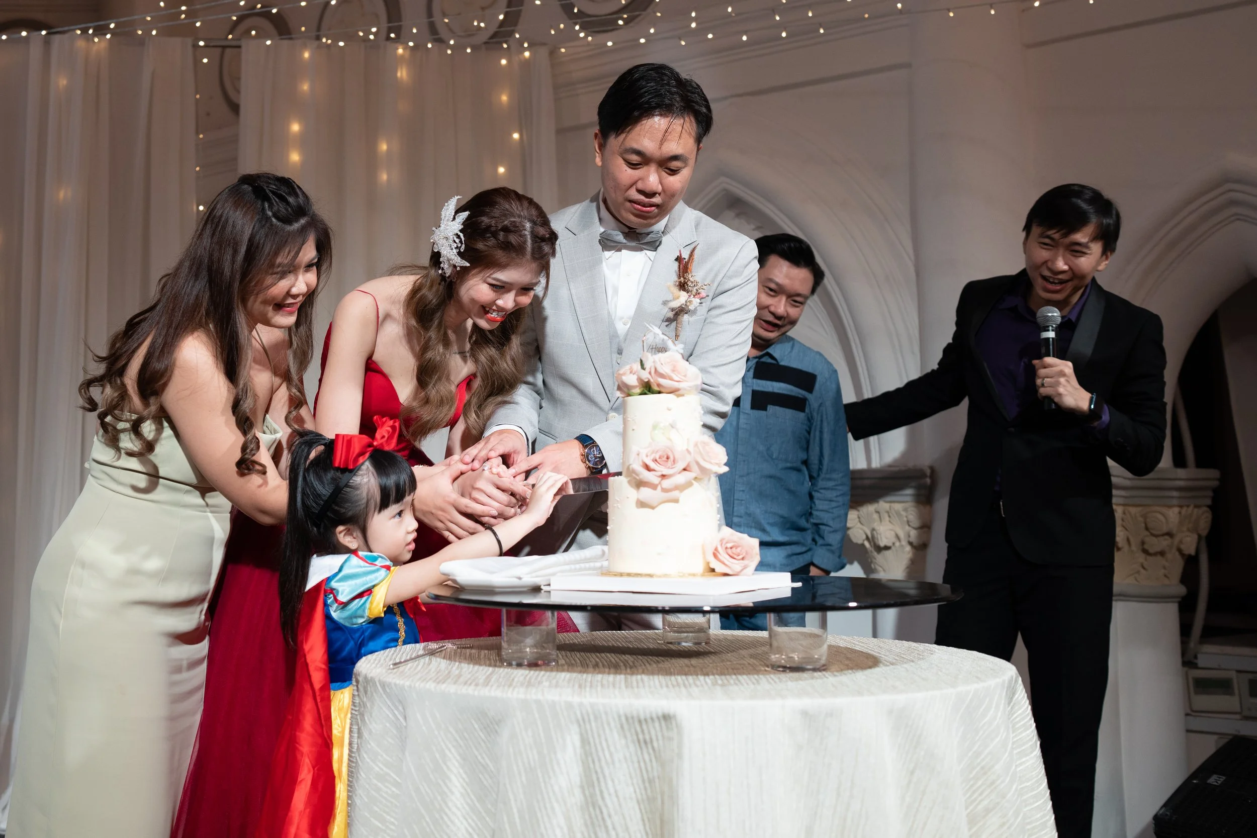 People cutting a wedding cake during a celebration, with a groom, bride, and guests gathered around.