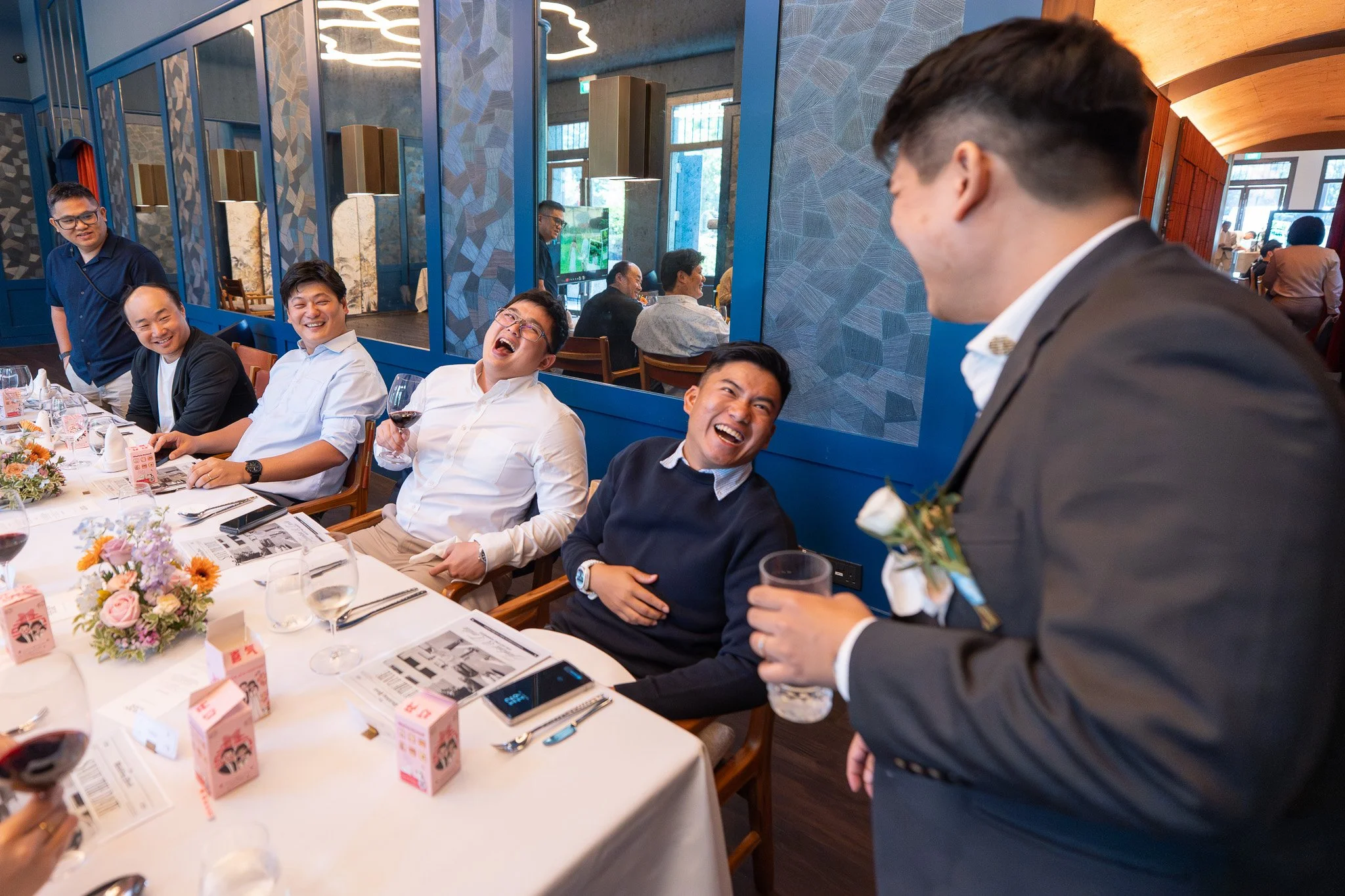 Group of men seated at a dining table, laughing and enjoying a celebratory toast with a man in a suit standing and smiling, in a restaurant with blue decor and flowers on the table.