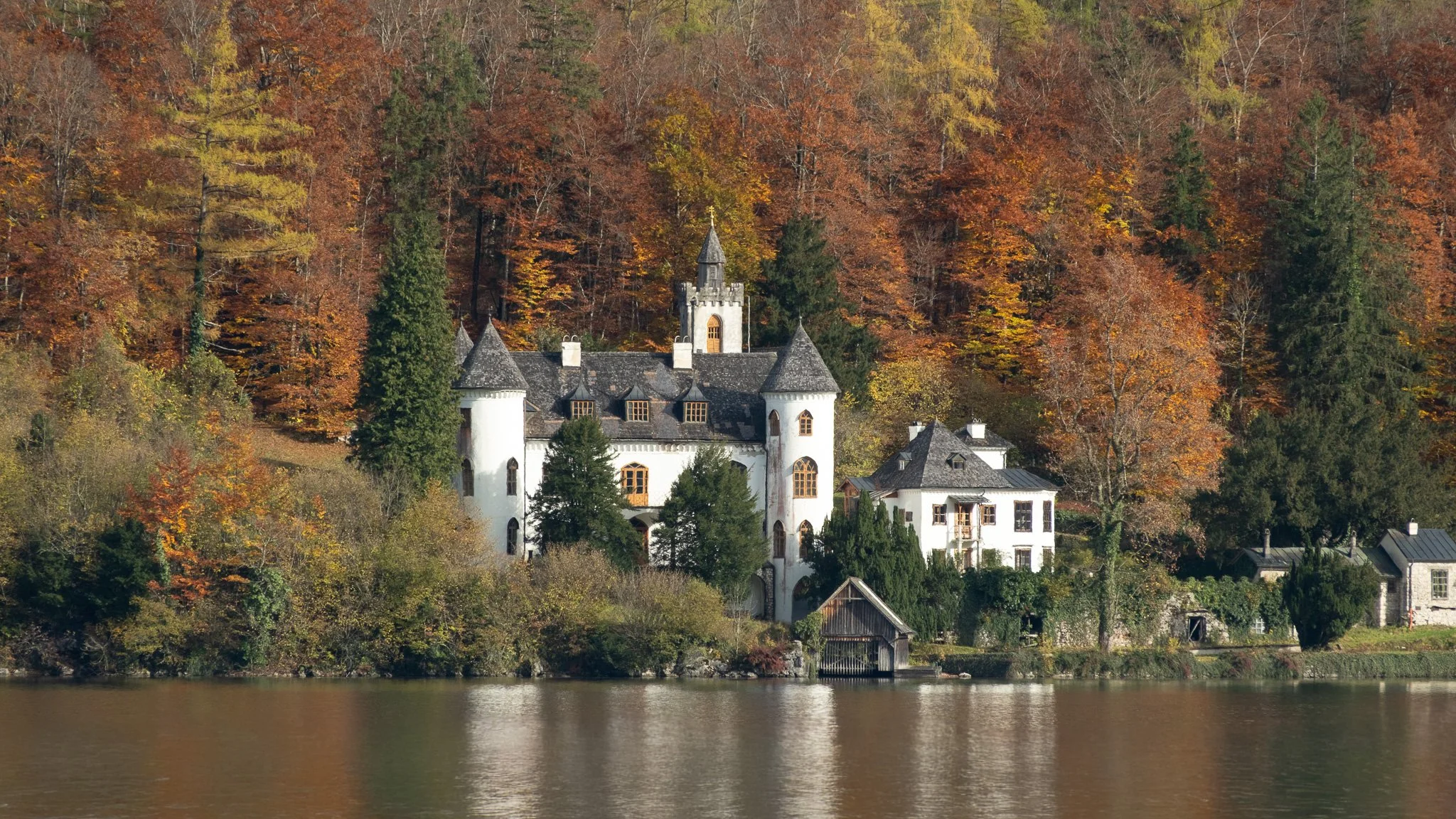A castle with white walls and dark roofs situated on a lakeshore, surrounded by autumn-colored trees and hillside.