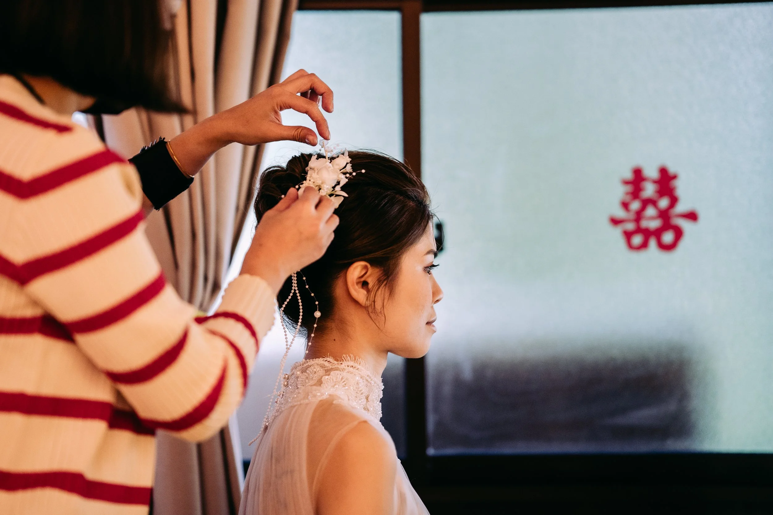 A woman is having her hair decorated with white floral accessories by another woman, in front of a window with beige curtains, with a red double happiness symbol on a gray wall in the background.