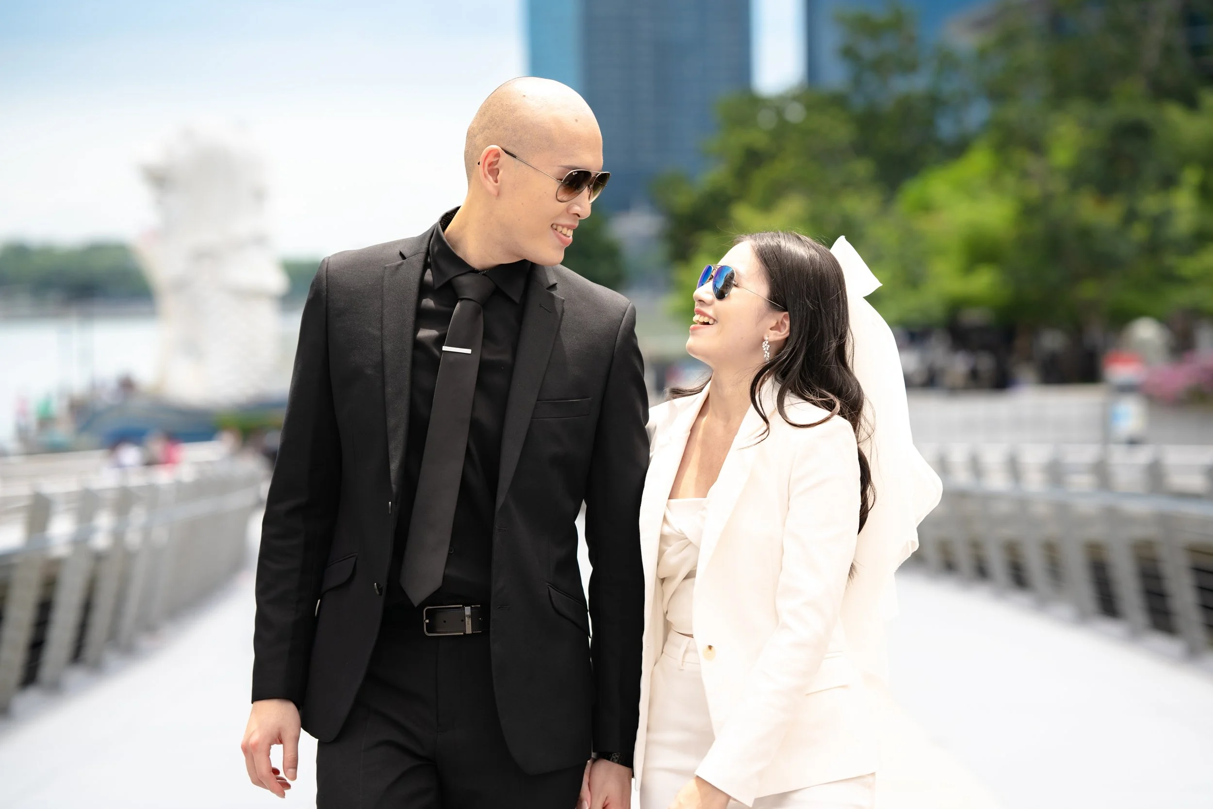 A couple dressed in formal attire walking outdoors, holding hands and smiling at each other.