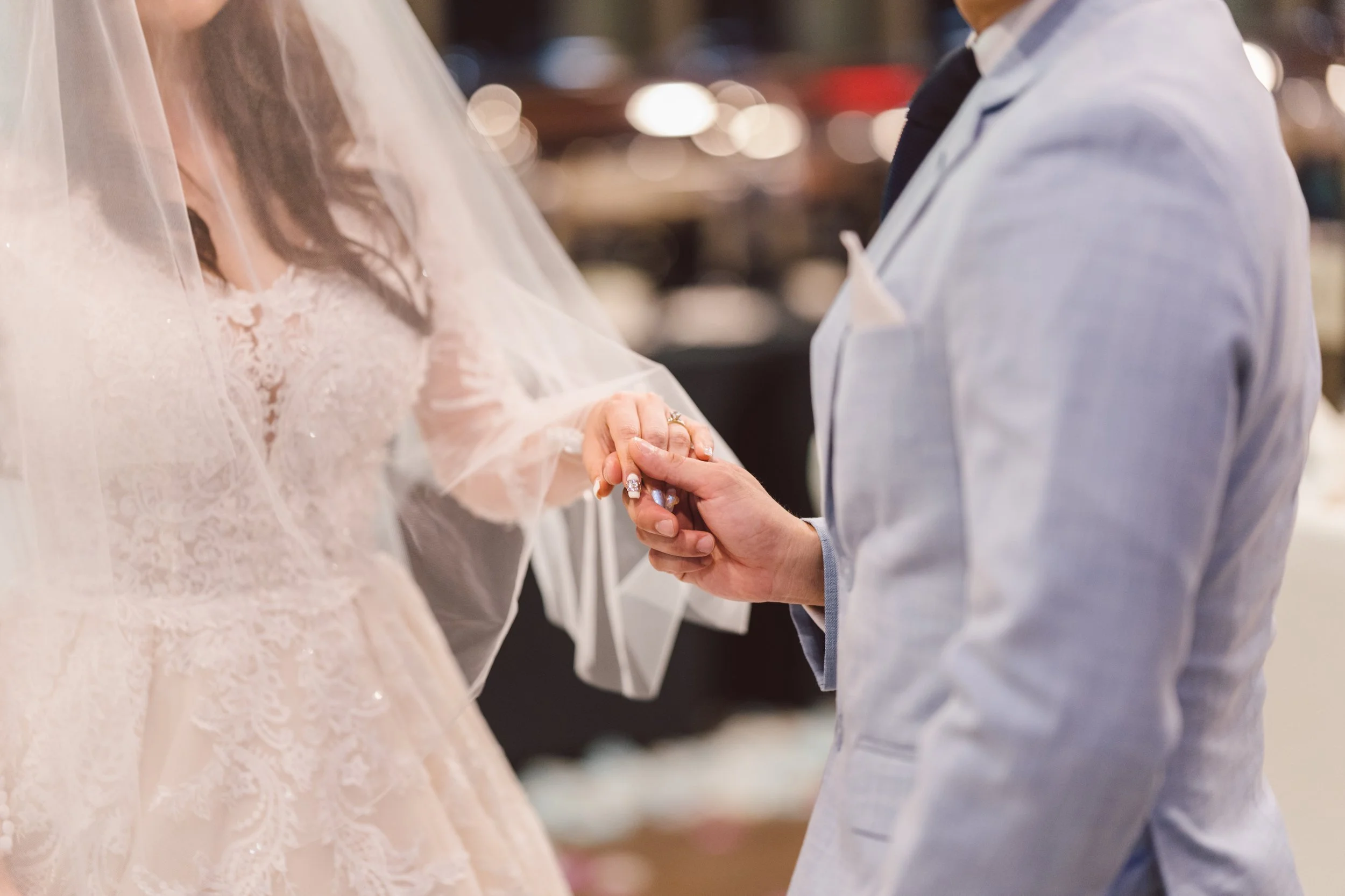 Close-up of a bride and groom holding hands during a wedding ceremony, with the bride wearing a lace wedding dress and veil, and the groom in a light gray suit.