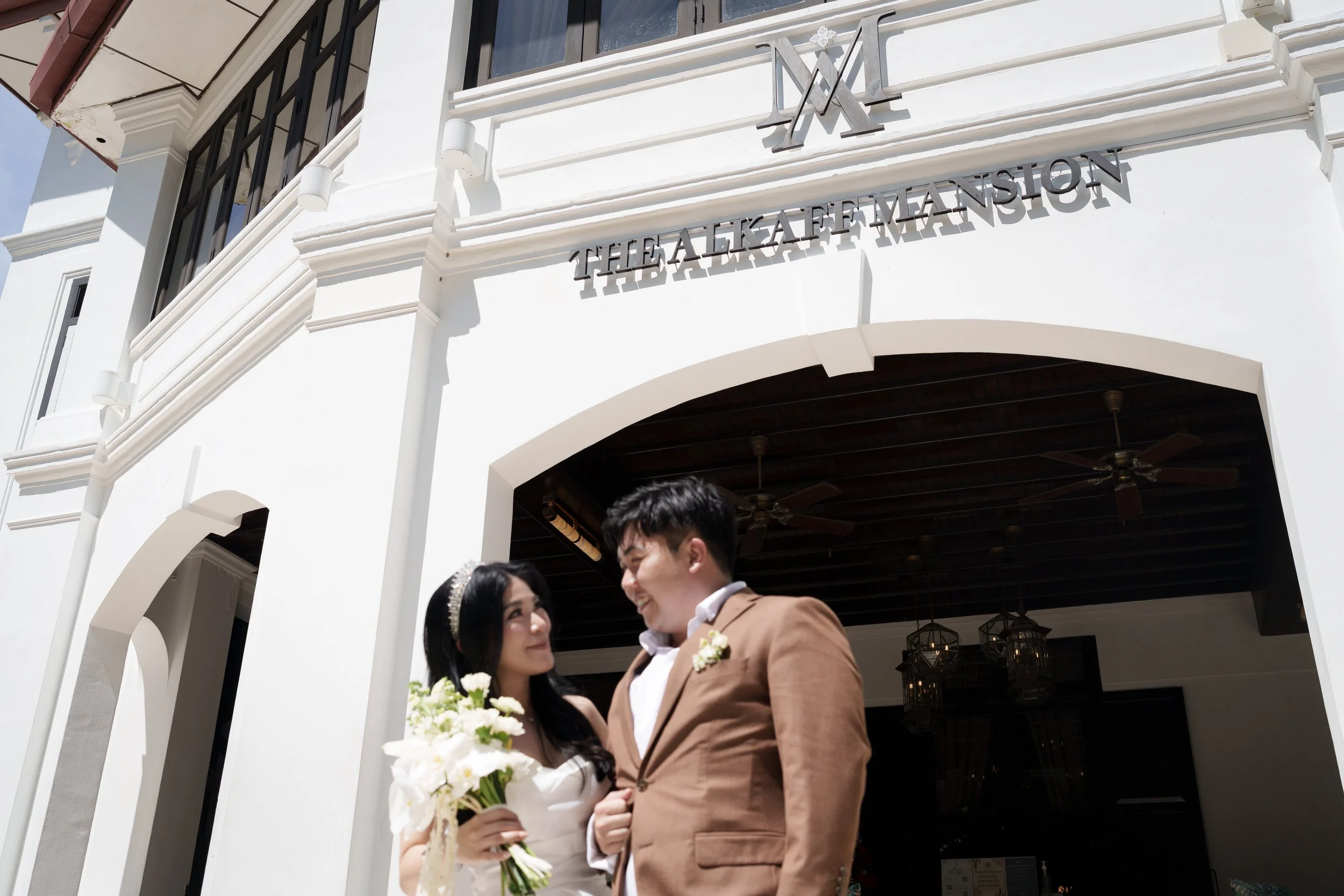 A wedding couple standing outside a building with a sign that reads 'The Alkaff Mansion'. The bride holds a bouquet of white flowers, and the groom wears a brown suit. They are smiling at each other.