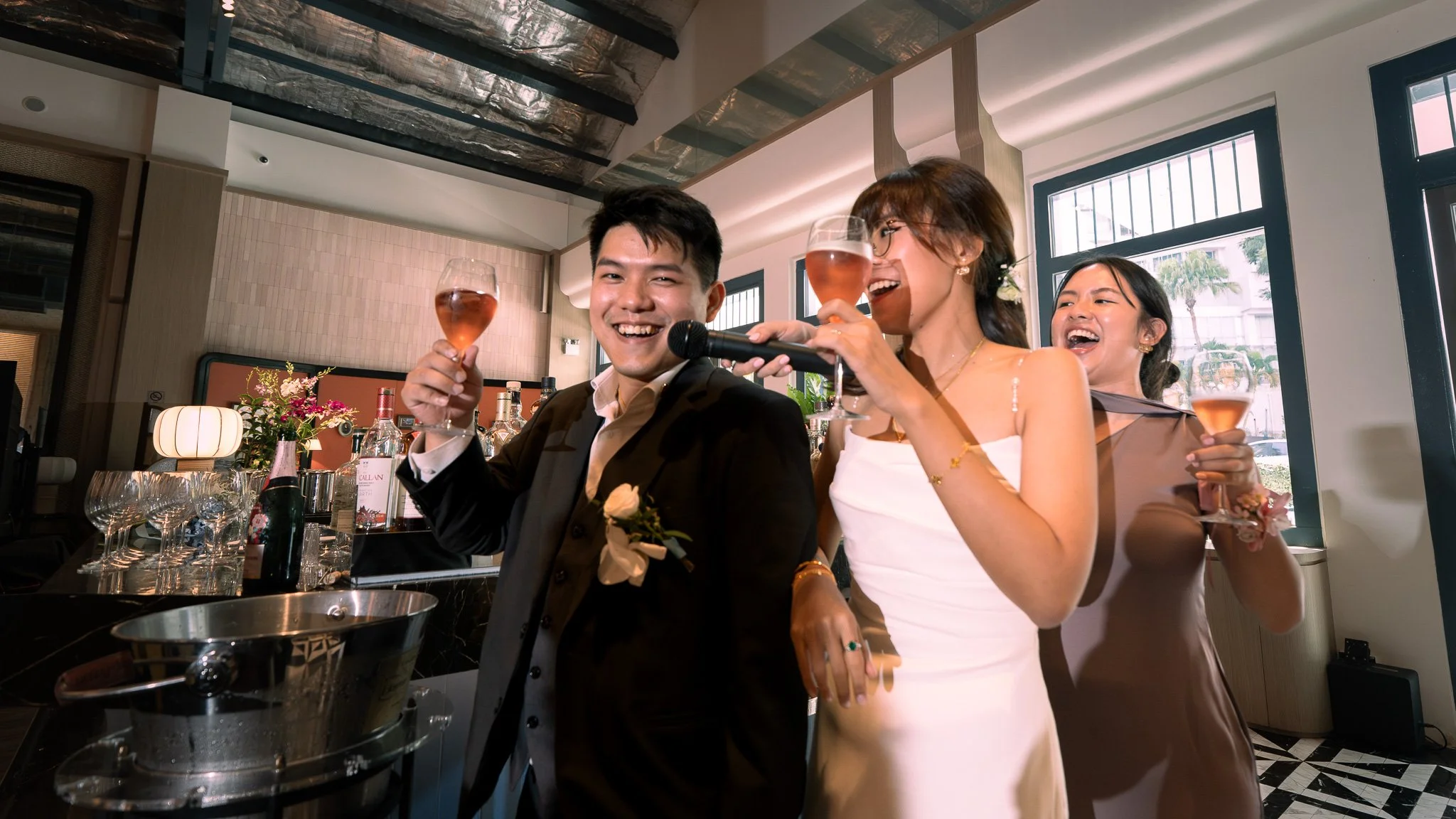 Three people celebrating with glasses of rosé wine at a decorated banquet table in a modern indoor venue, smiling and enjoying the moment.