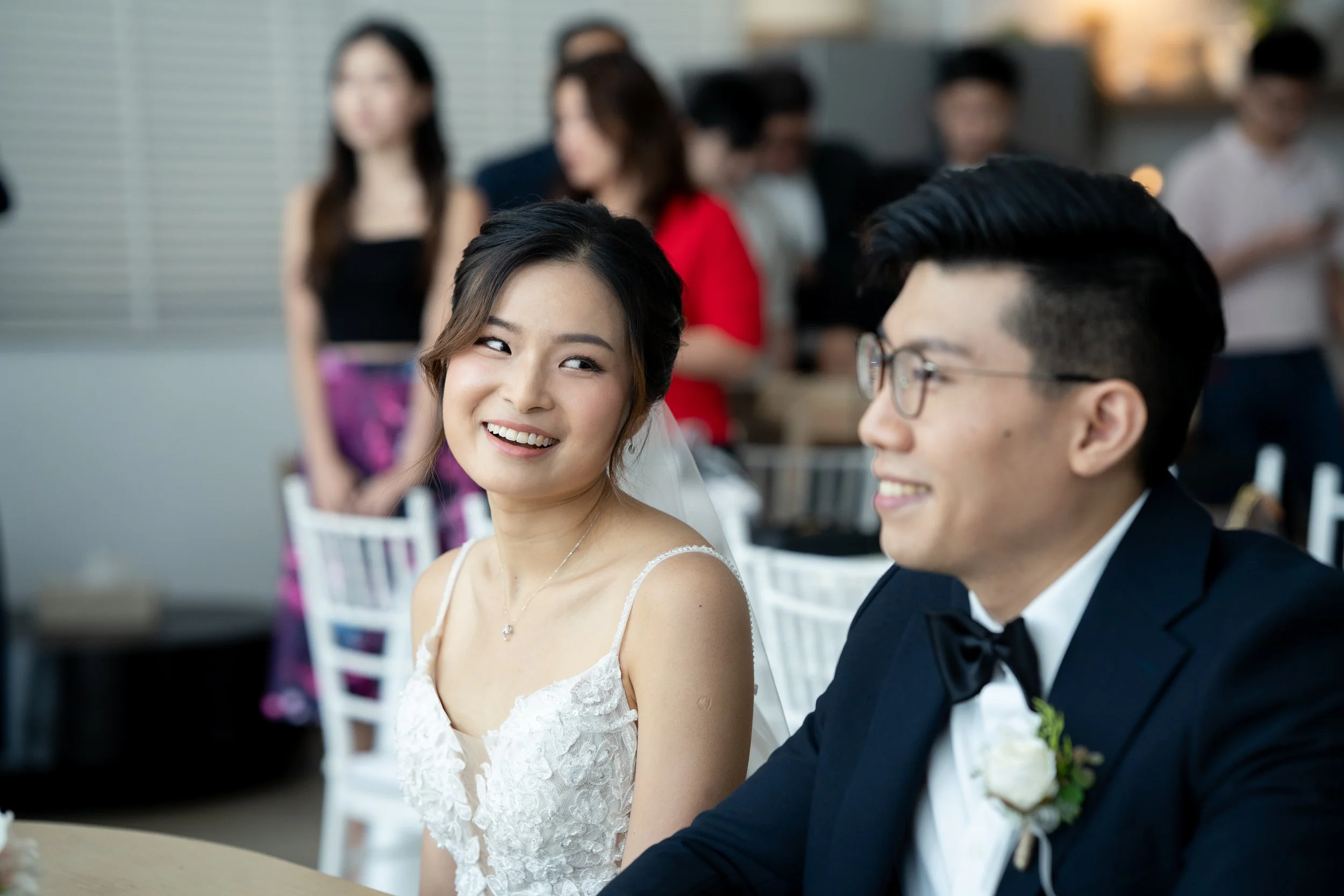 A smiling bride and groom sit at a reception, with other guests in the background.
