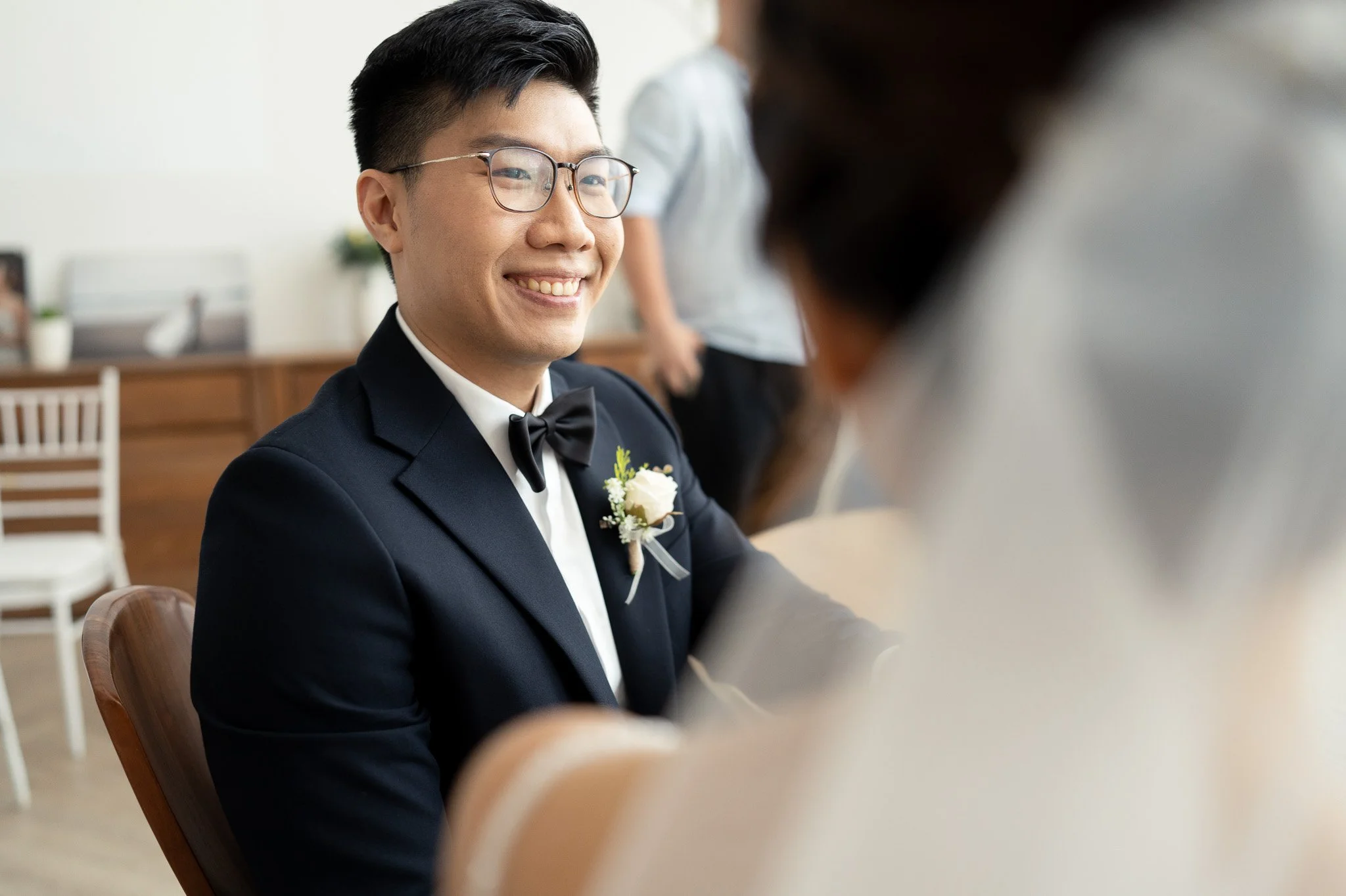 A man in a tuxedo with glasses and a boutonniere, smiling at someone across a table during a wedding or formal event.
