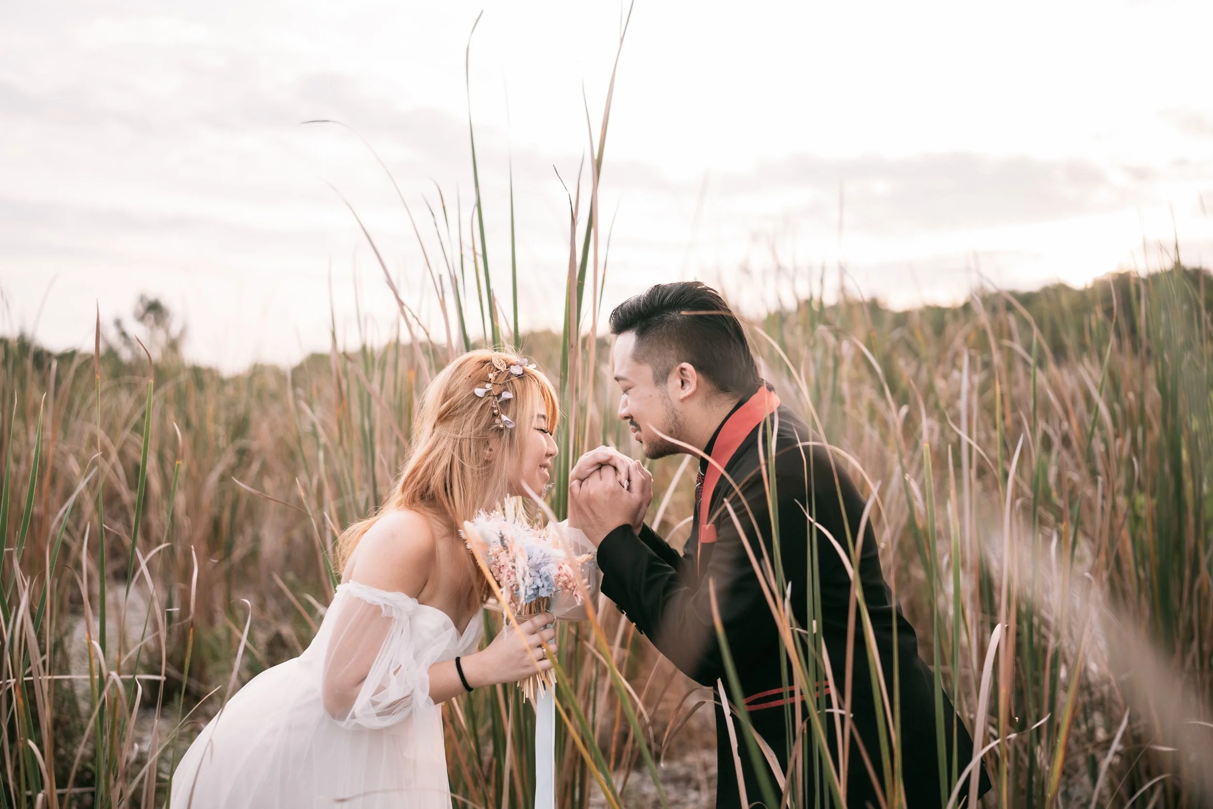 A couple in wedding attire in a field, holding hands and smiling at each other.
