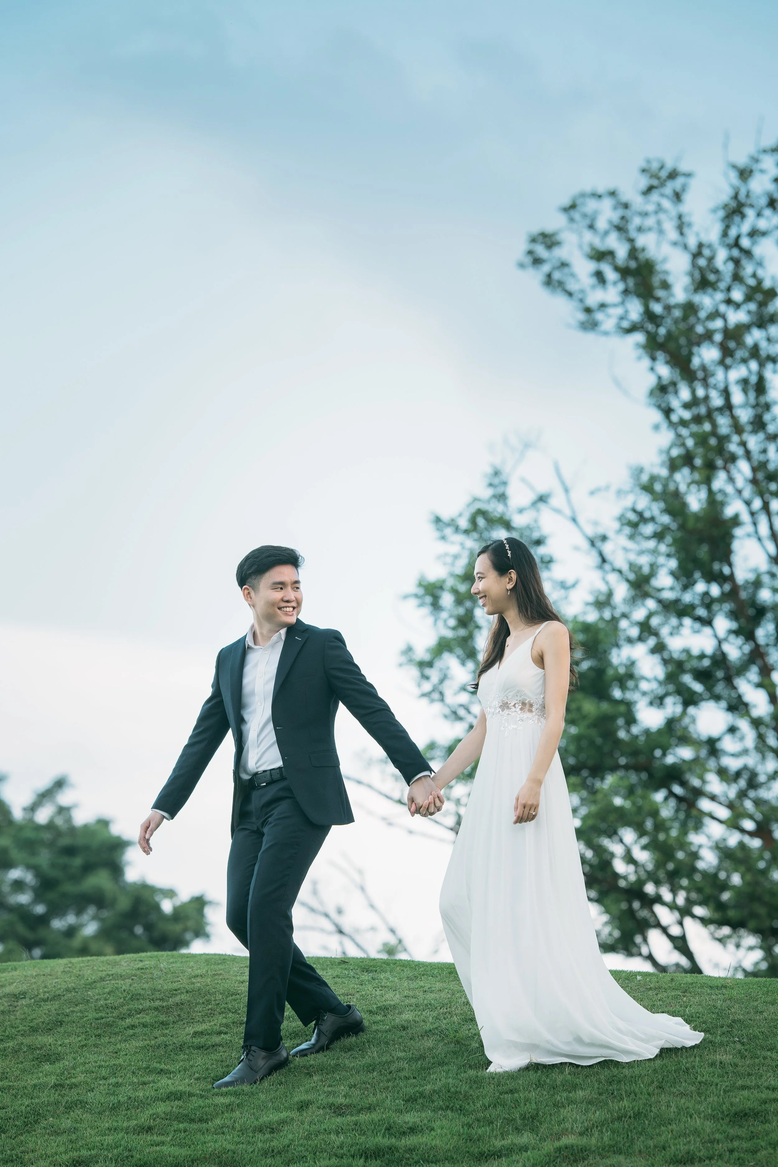 A couple in wedding attire holding hands and walking on grass outdoors, smiling at each other with trees and a blue sky in the background.