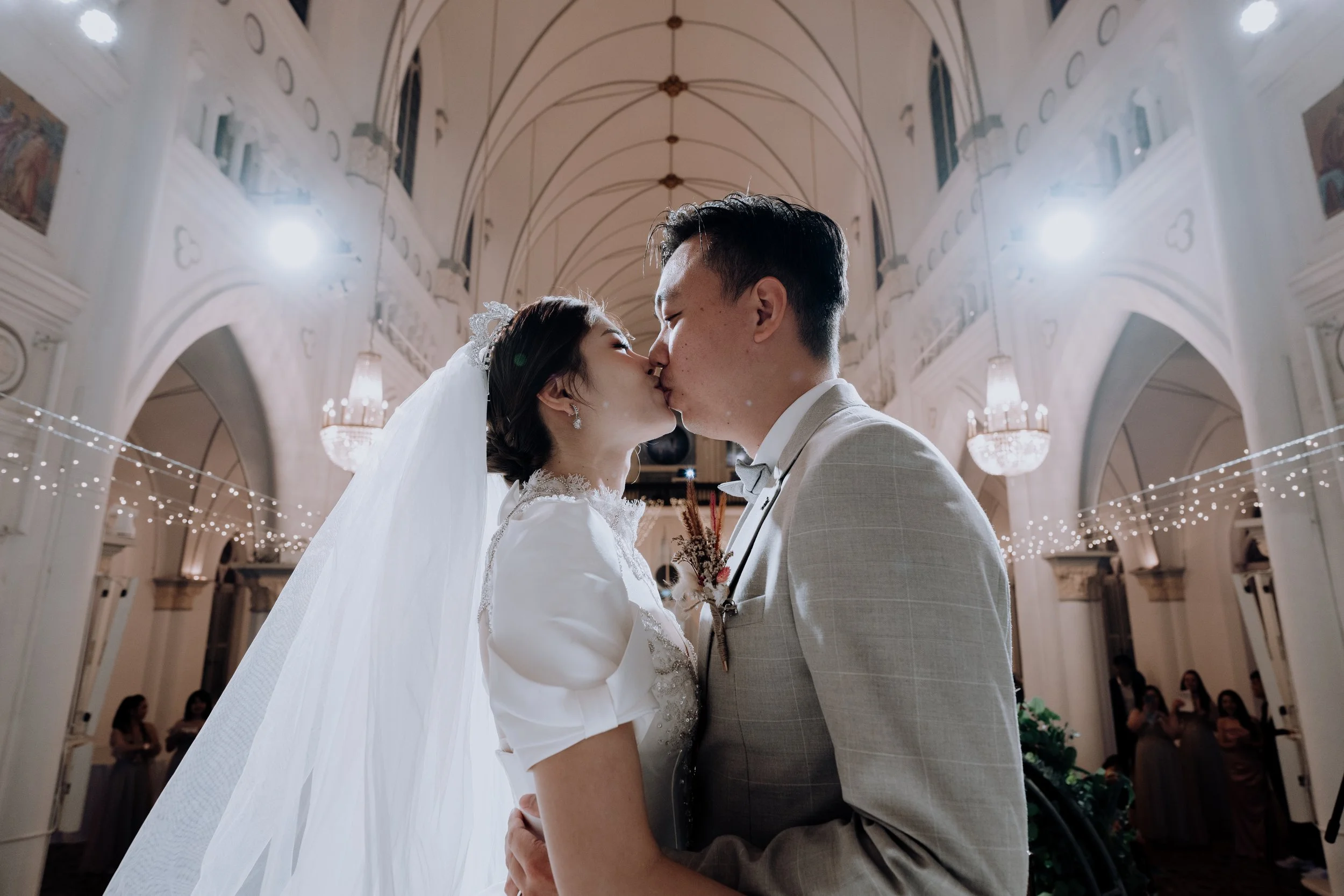 A bride and groom sharing a kiss during their wedding ceremony inside a decorated church with high arched ceilings and chandeliers.