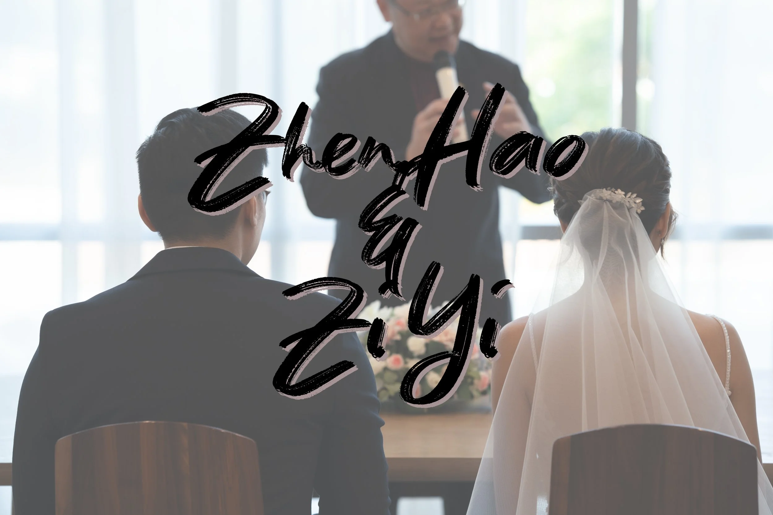 A wedding ceremony with a bride and groom sitting at a table, facing an officiant. The couple is dressed in formal attire; the bride in a white wedding dress with a veil and the groom in a dark suit. The scene is indoors with natural light coming thr