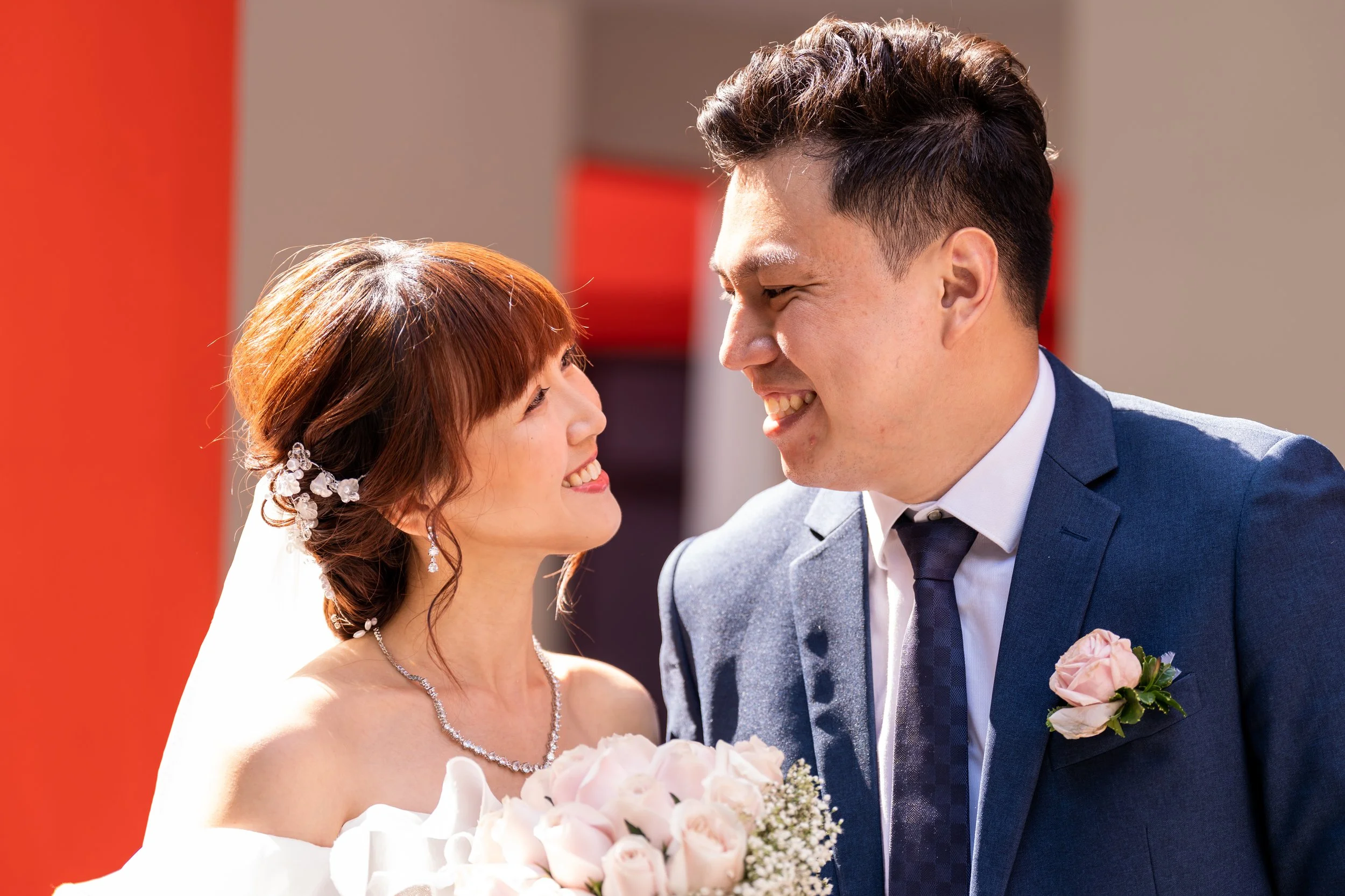 A newlywed couple smiling at each other outdoors. The bride has red hair styled in loose waves with a floral hairpiece, wearing a white off-the-shoulder wedding gown, jewelry, and holding a bouquet of pink roses. The groom has short dark hair, wearin