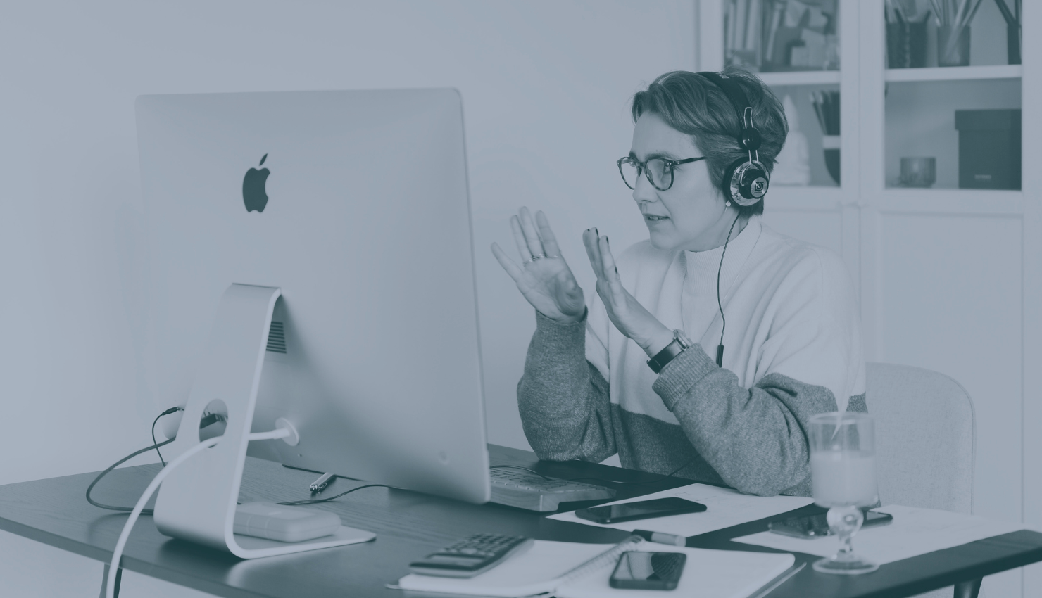 A woman with glasses and headphones sitting at a desk, gesturing with her hands while speaking on a video call on her computer.