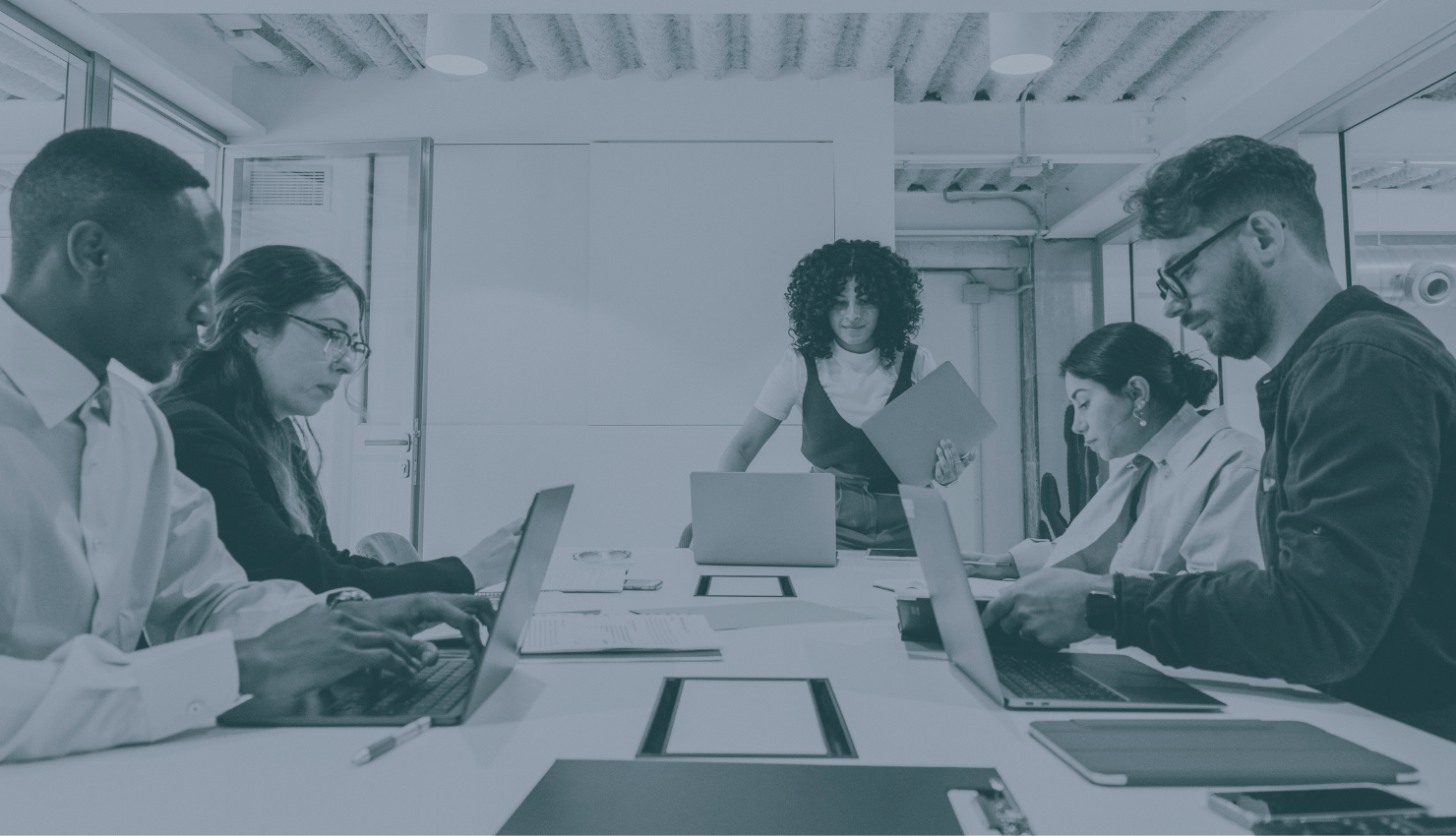 A diverse group of five people in a professional office conference room working on laptops and reviewing documents, with one woman standing and holding a folder in the background.