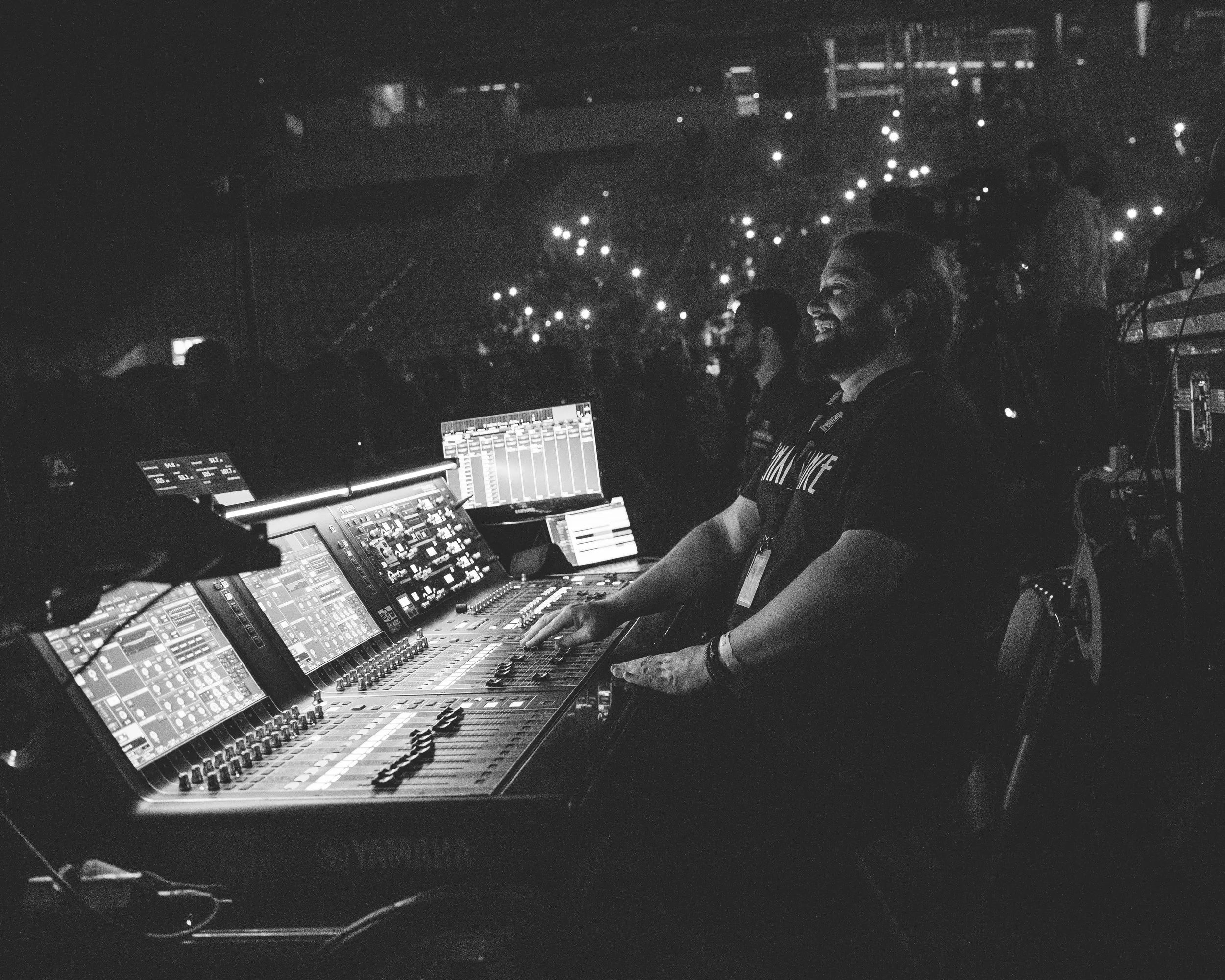 Persona sonriendo en un puesto de control de audio en un concierto, con pantallas y controles en la mesa, en un ambiente oscuro con luces en el fondo.