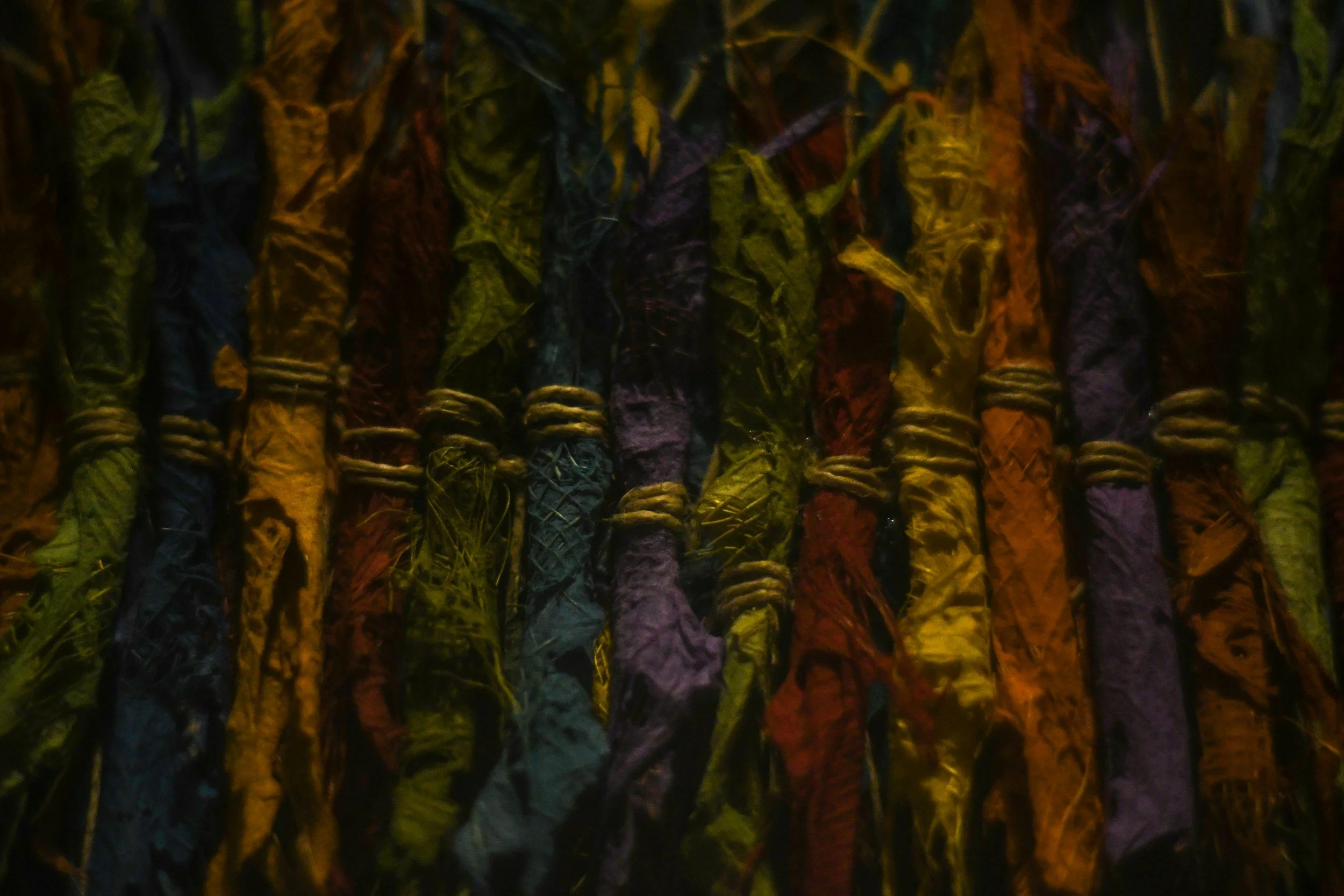 A close-up of various colorful cloths or fabrics tied around sticks or poles, creating a textured pattern with yellow, purple, red, green, and black colors.