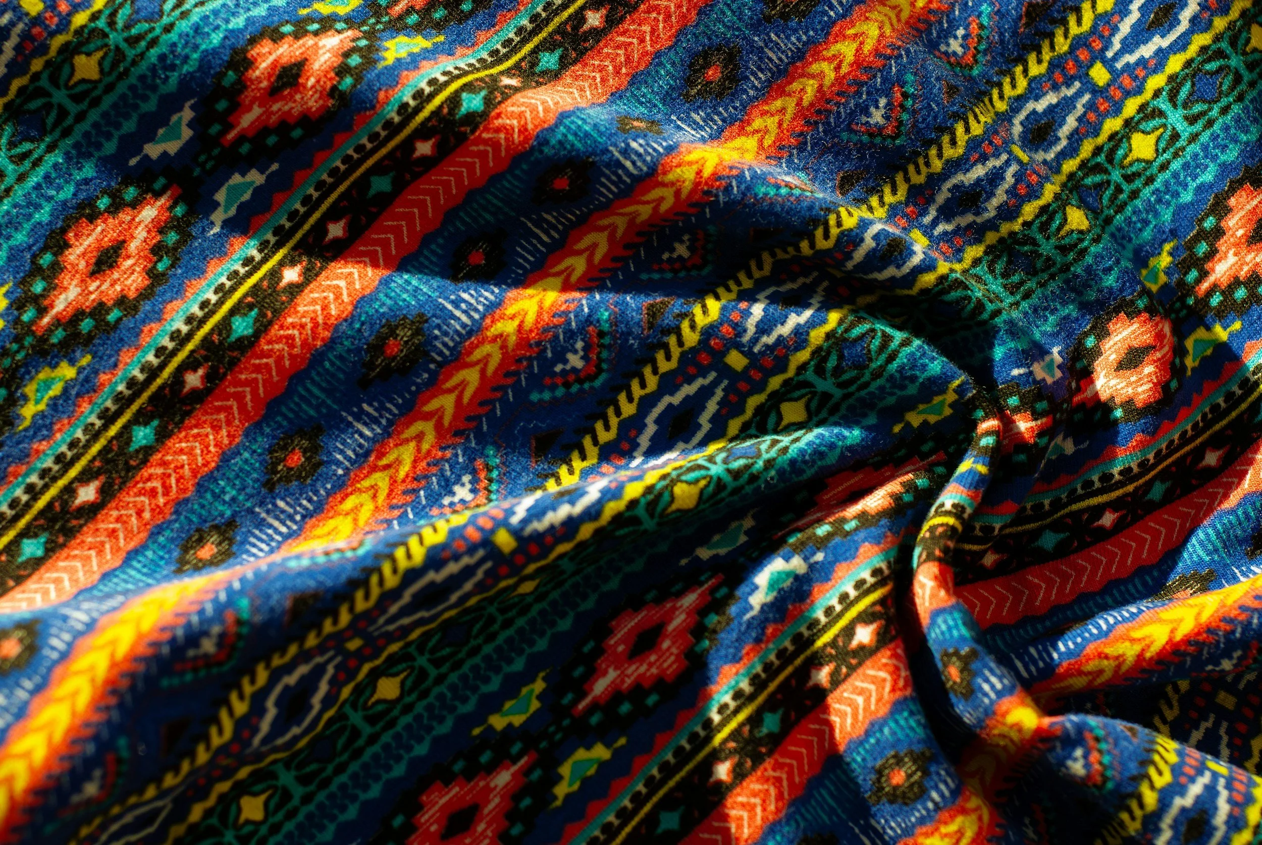 Colorful, patterned fabric with intricate designs in red, blue, yellow, green, and black, draped and slightly wrinkled.