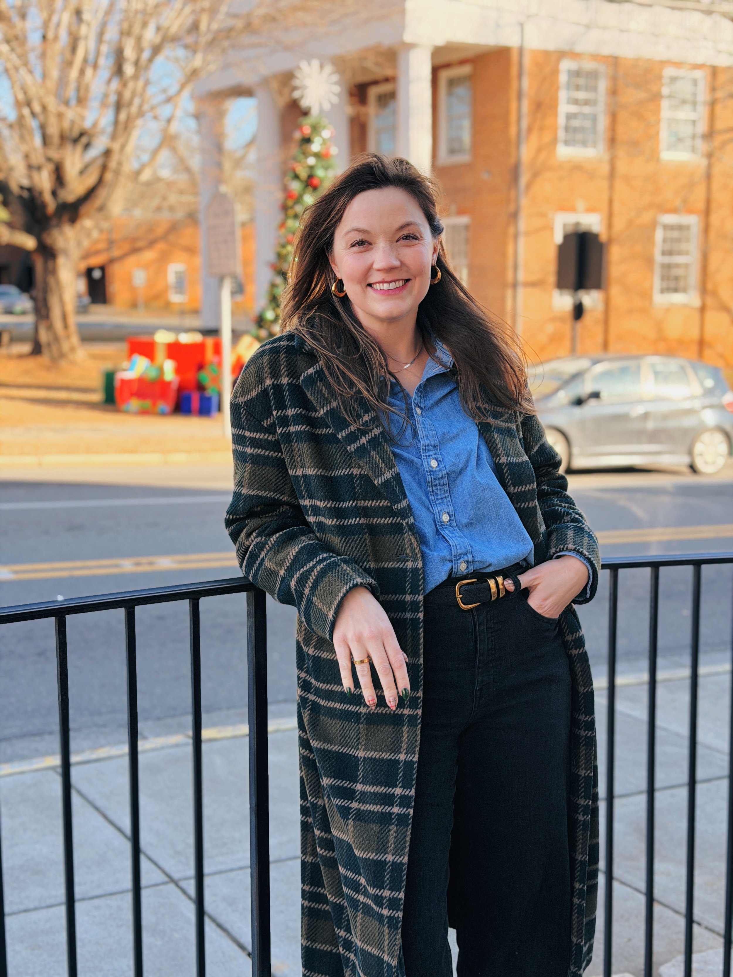 A woman standing outdoors in front of a railing, smiling, during daytime with a decorated Christmas tree and gift boxes in the background.
