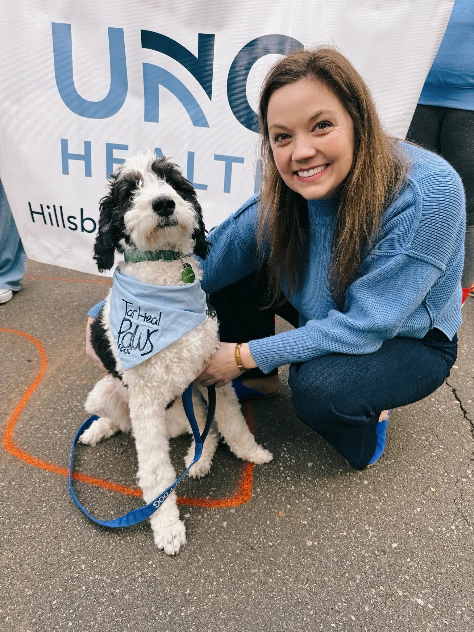 A woman crouching next to a black and white dog therapy dog with a blue bandana at a health event. There is a banner behind them with UNC Health
