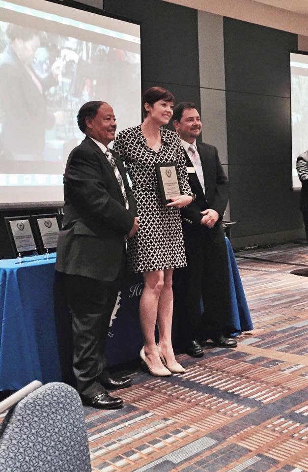 A woman in a black and white patterned dress holding an award, standing between two men in business suits, at an award ceremony in a conference room.