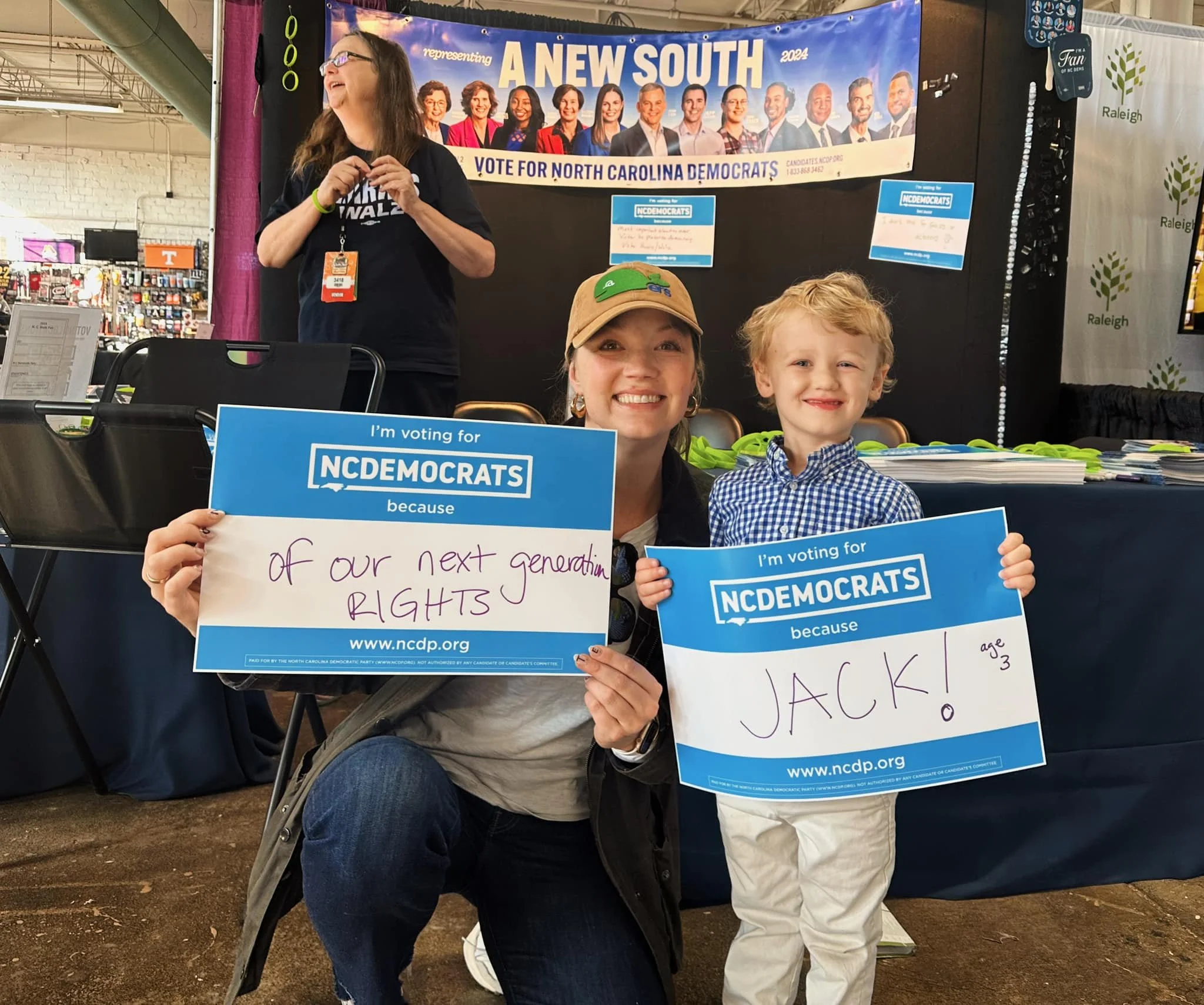 A woman and a young boy at a political event holding signs supporting the NC Democrats, with a banner for a South Carolina candidate in the background.