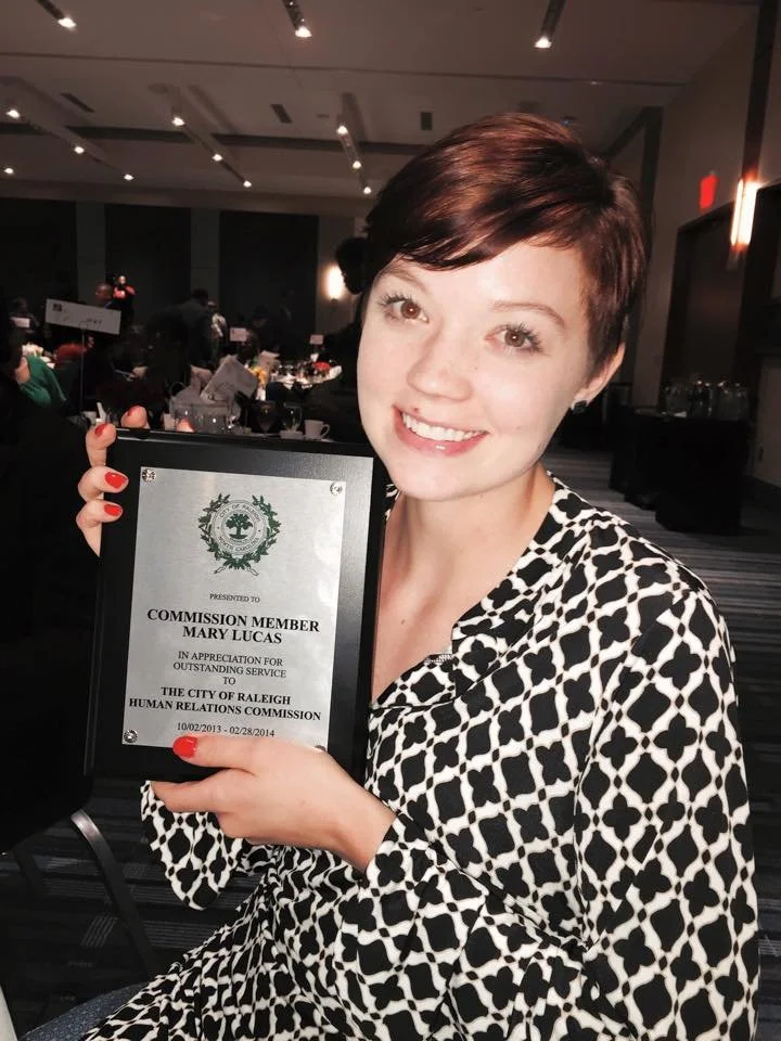 A young woman with short brown hair and a bright smile holding a plaque at an indoor event or awards ceremony. She is wearing a black and white patterned dress and has red nail polish.