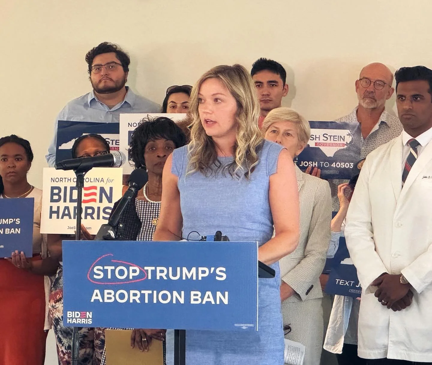 A woman in a blue dress speaking at a political rally with a blue sign that reads "STOP TRUMP'S ABORTION BAN." Several people stand behind her, some holding signs supporting Joe Biden and Kamala Harris, and others listening attentively.