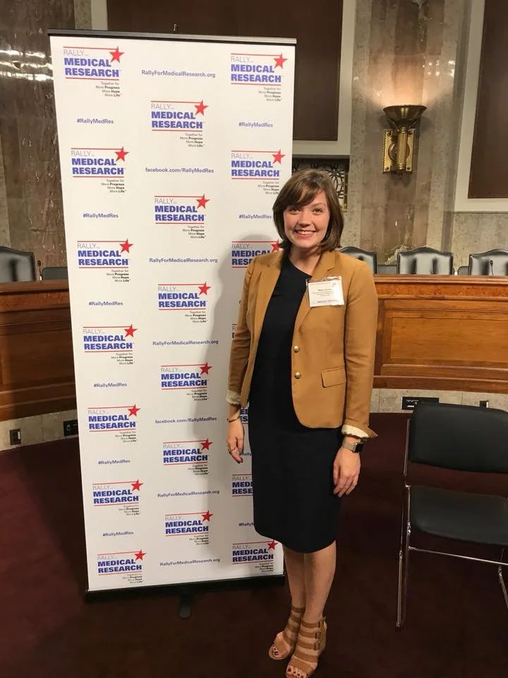 A woman standing in front of a Rally for Medical Research banner, smiling, wearing a tan blazer, black dress, and beige high-heeled sandals.