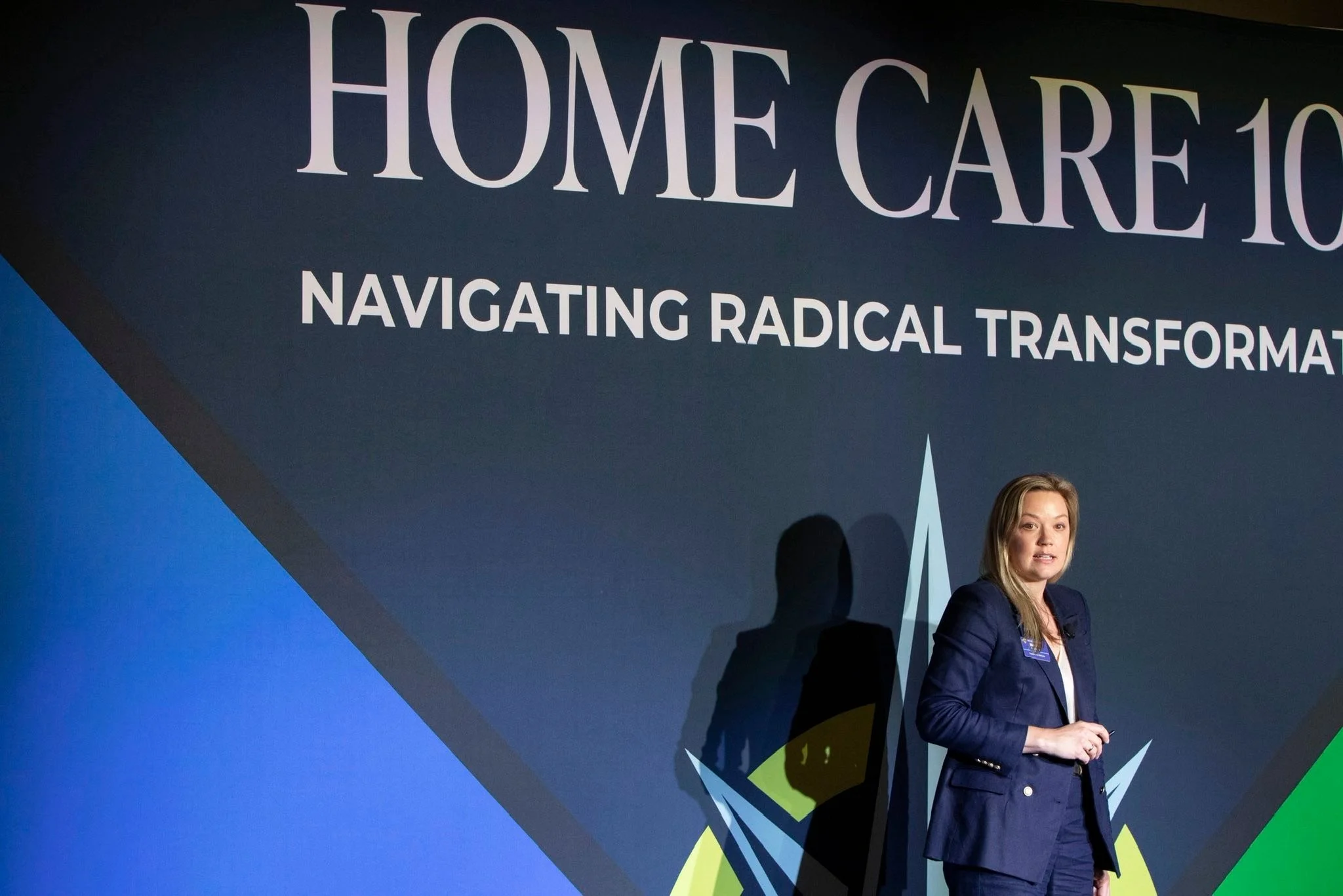 Woman in navy blazer speaking at a conference on home care, with a dark backdrop displaying the text 'HOME CARE 10' and 'NAVIGATING RADICAL TRANSFORMATION'.