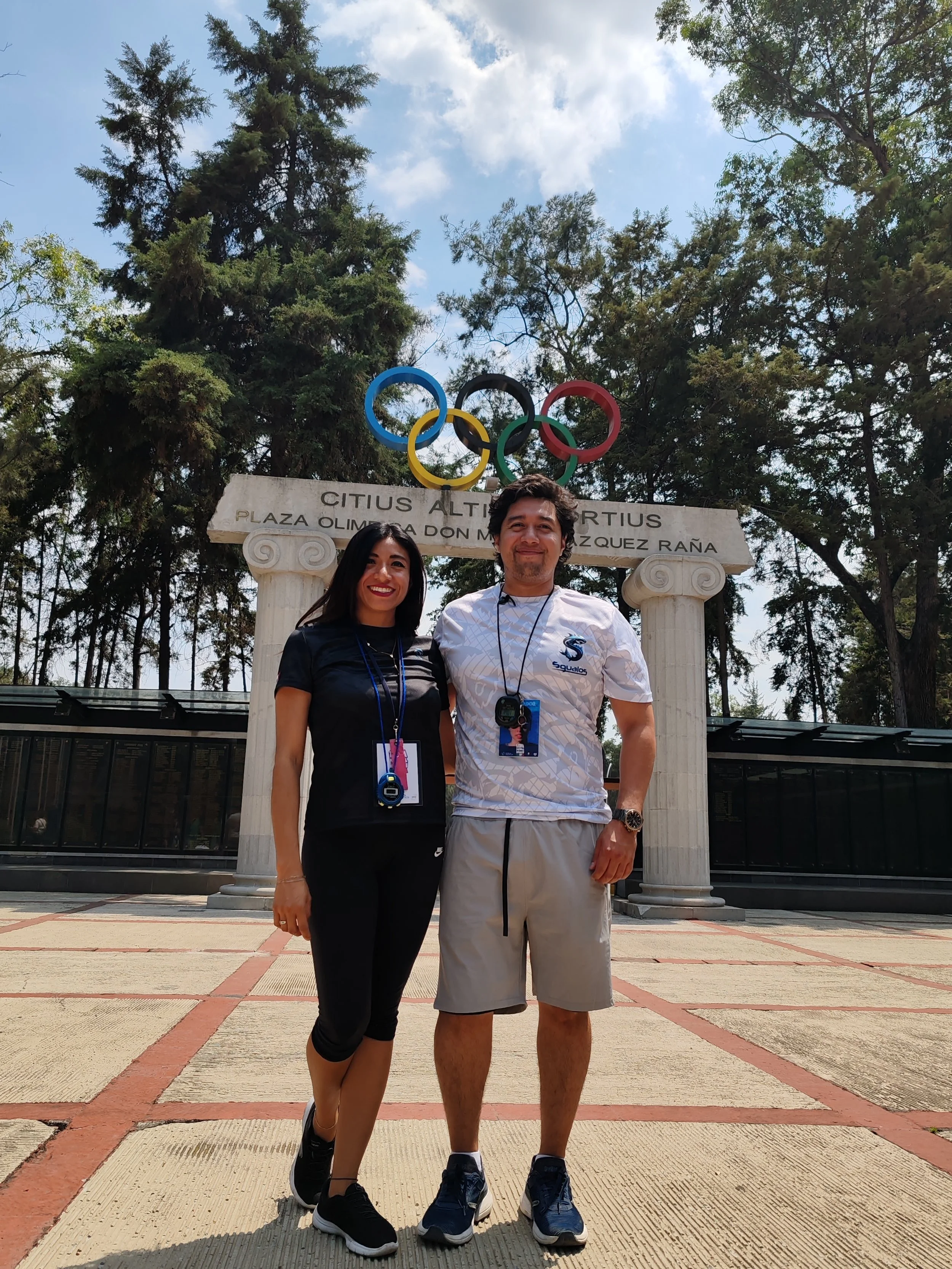Joel Espinoza y Mariana Franco entrenadores de natacion profesionales en primer plano, en el nacional infantil, con equipos de cronometraje colgados del cuello, posando en el comite olimpico mexicano con los anillos olímpicos.