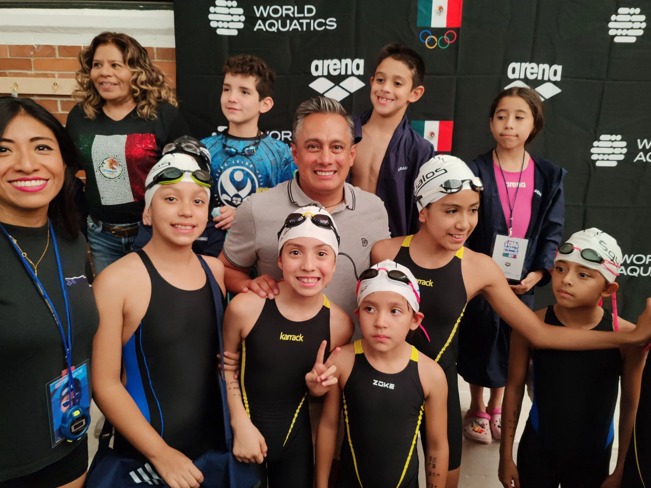 Grupo de niños en una competencia de natación, nacional infantil en el comite olmipico mexicano con Fernando Platas exclavadista olimpico, frente a un fondo con logos de World Aquatics, Arena y la bandera de México.