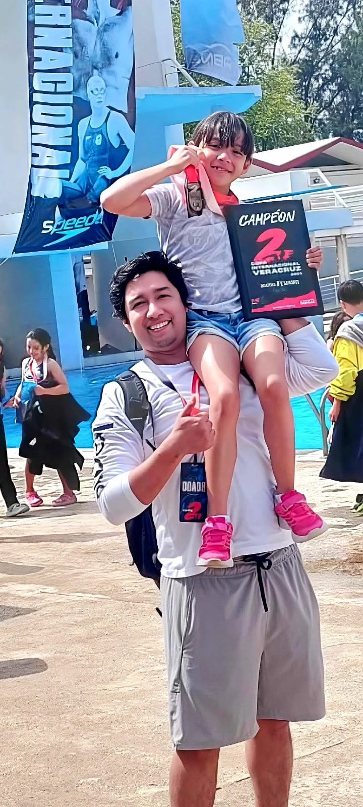 El entrenador Joel Espinoza lleva a Fernanada Anta en los hombros en una celebración del campeonato de natacion, la copa internacional, la niña sostiene un cartel de campeón de una competencia en Veracruz, en un evento al aire libre.