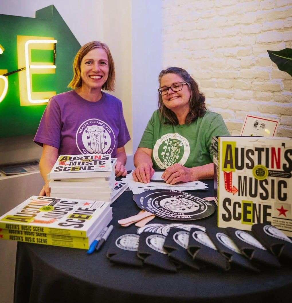 Two women smiling at a table with books, pamphlets, and promotional materials promoting 'Austins Music Scene.' They are wearing purple and green shirts with a logo that says 'Health Alliance for Austin Musicians.' The background includes a neon sign and a brick wall.
