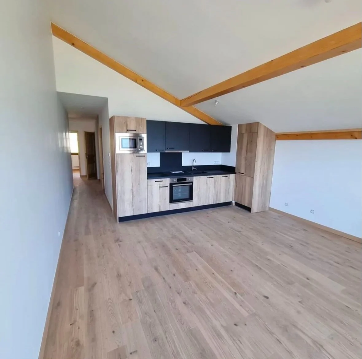 Une cuisine dans un appartement avec un plafond en pente, des poutres en bois apparentes, des armoires en bois clair, un plan de travail noir, un four encastré et un micro-ondes au mur.