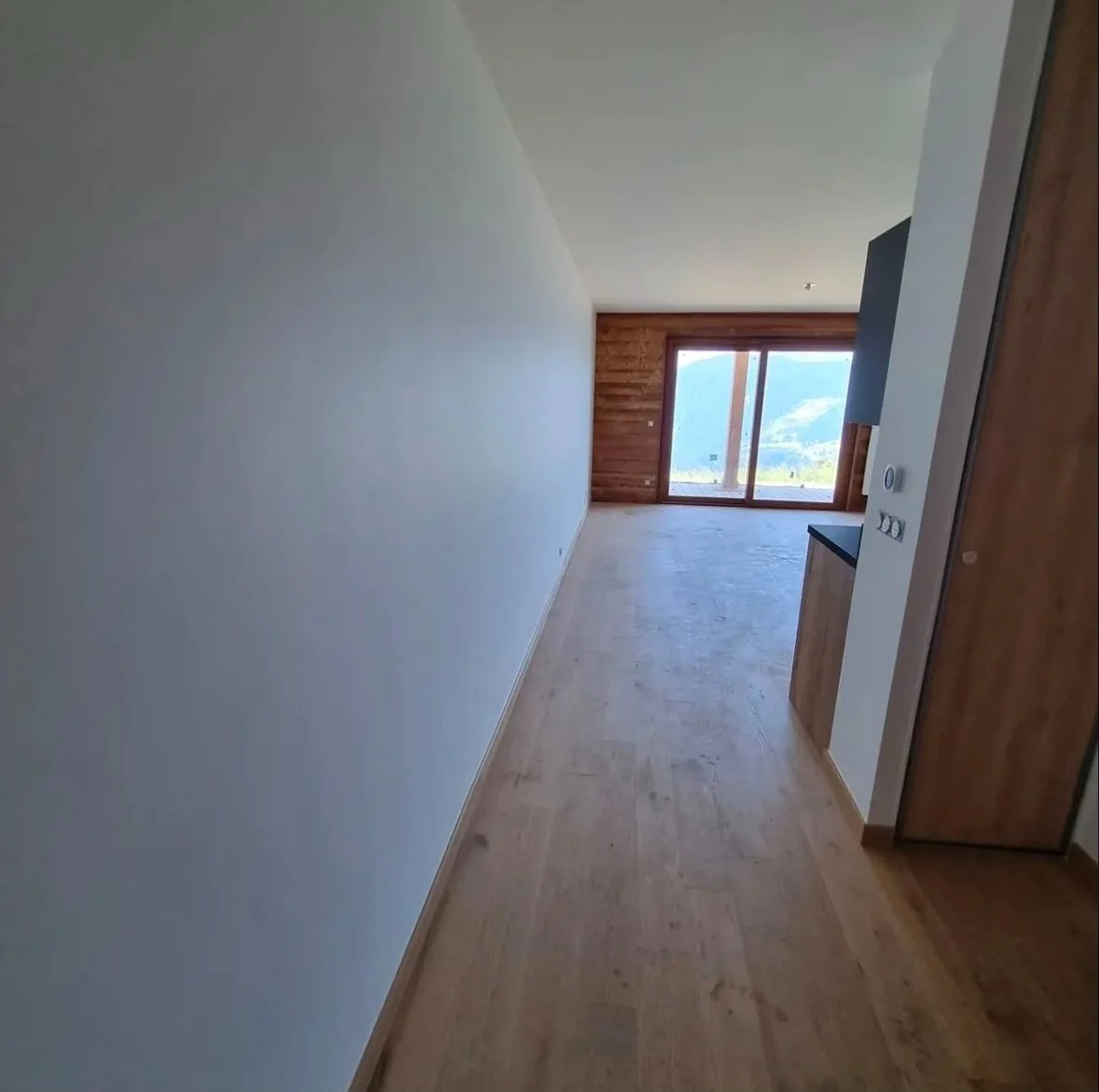 Intérieur d’un appartement vide avec sol en bois, murs blancs et un grand balcon avec vue sur la montagne.
