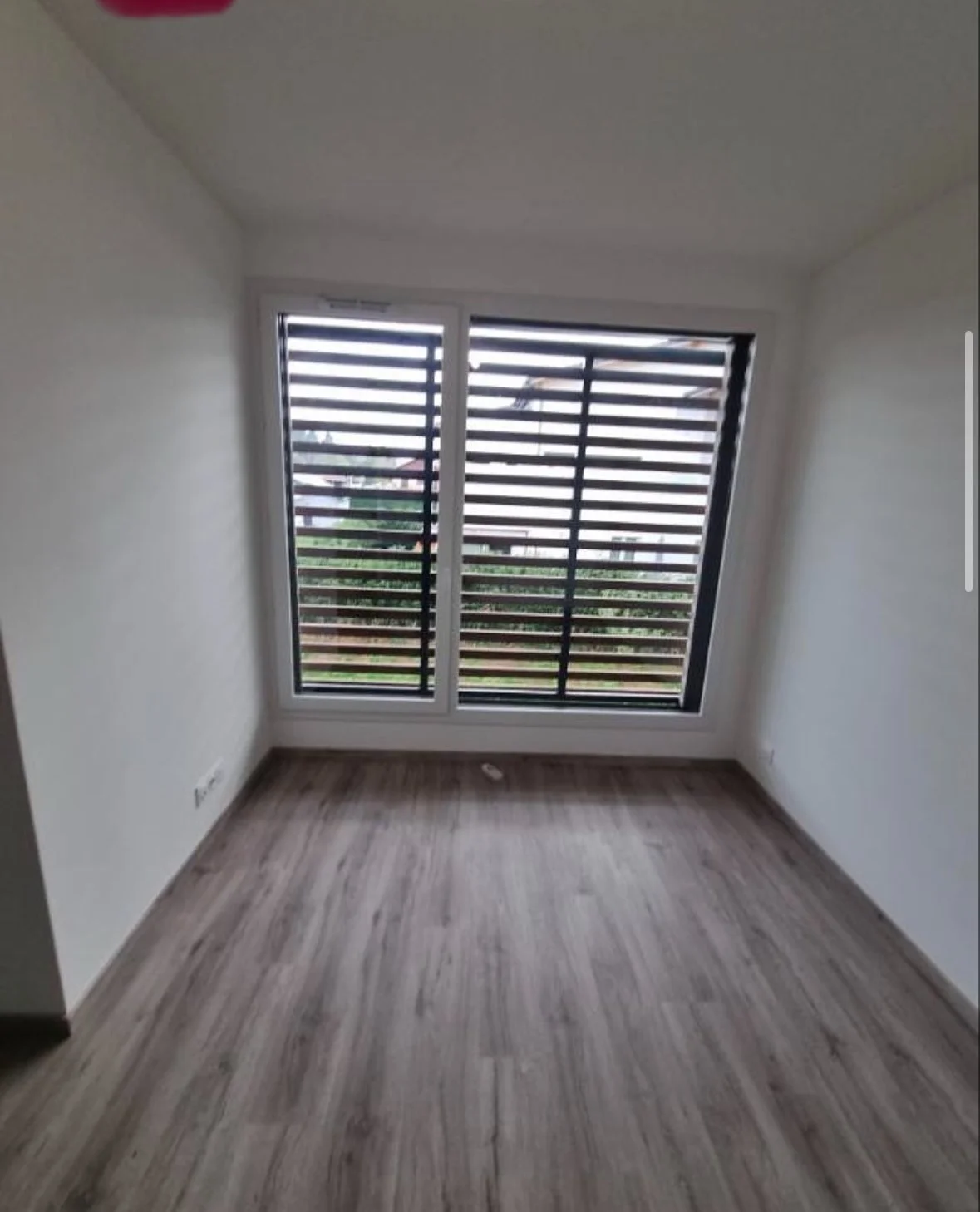 Une pièce vide avec un sol en bois clair, des murs blancs et une grande fenêtre avec des volets en bois.