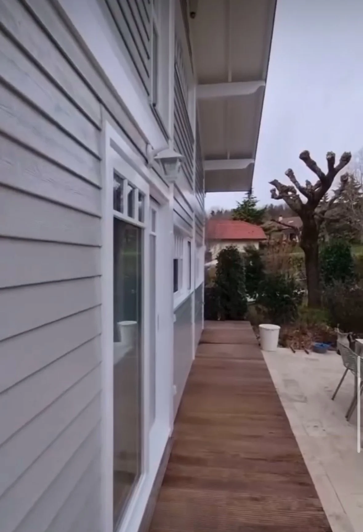 Balcon en bois à côté d'une maison avec des fenêtres en verre, arbre taillé et jardin avec des buissons et une chaise en métal.