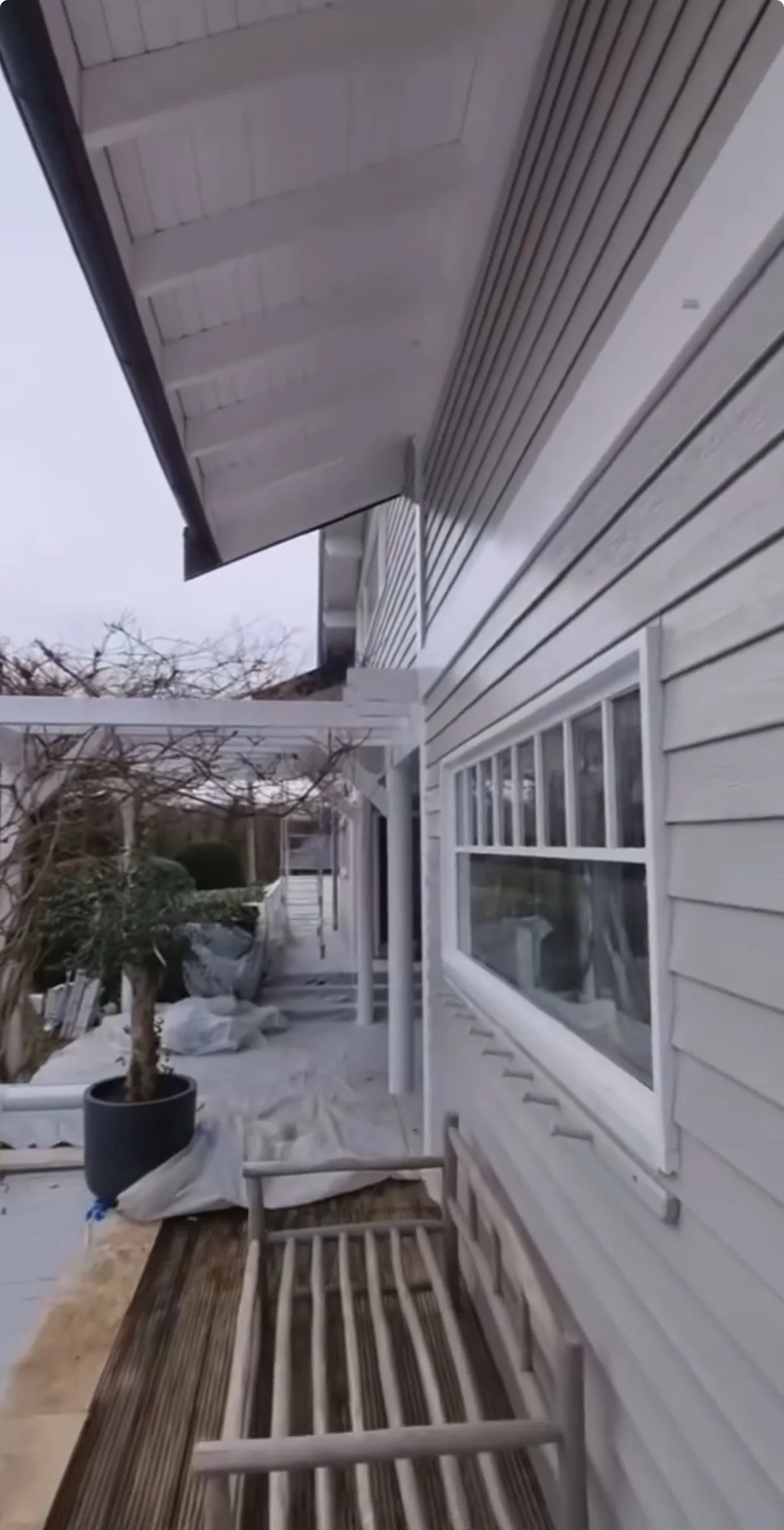 Vue d'une maison en bois blanc avec fenêtre, terrasse et mobilier extérieur, bordée de végétation, dans une journée nuageuse.