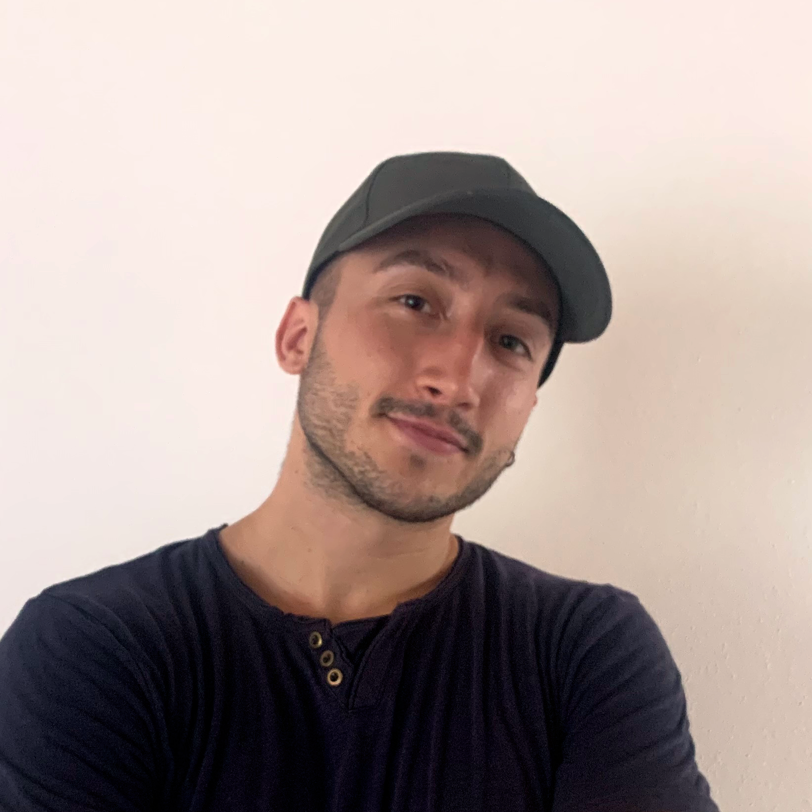 Ritratto di un uomo con cappellino e maglietta nera, sfondo semplice e chiaro.