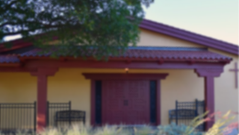 Exterior shot of St. Christopher's of Lubbock, a light brown brick church with dark red accents and red doors. There are a couple empty benches outside.