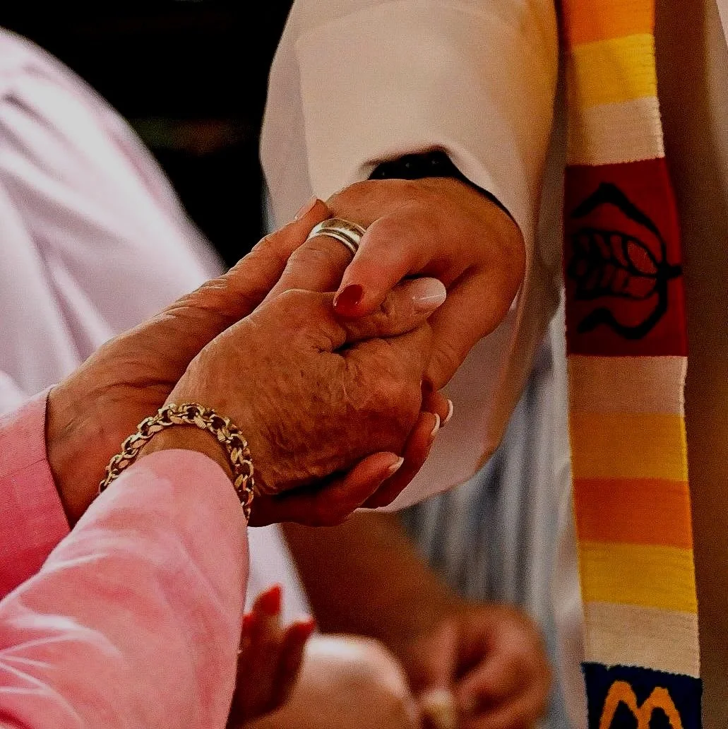 Two people holding hands, one appears elderly with jewelry, the other possibly a younger person wearing a pink shirt, close-up of interlocked hands.