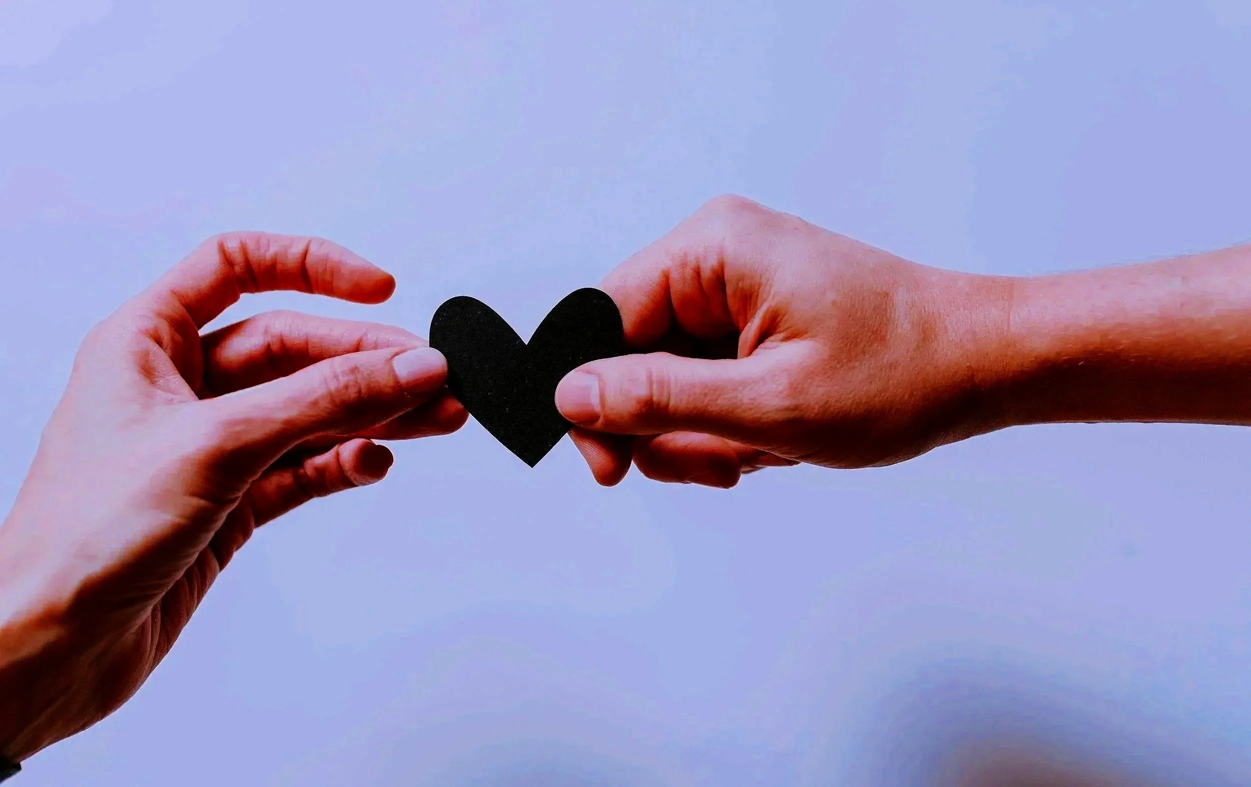 Two hands holding a black paper heart against a light blue background.