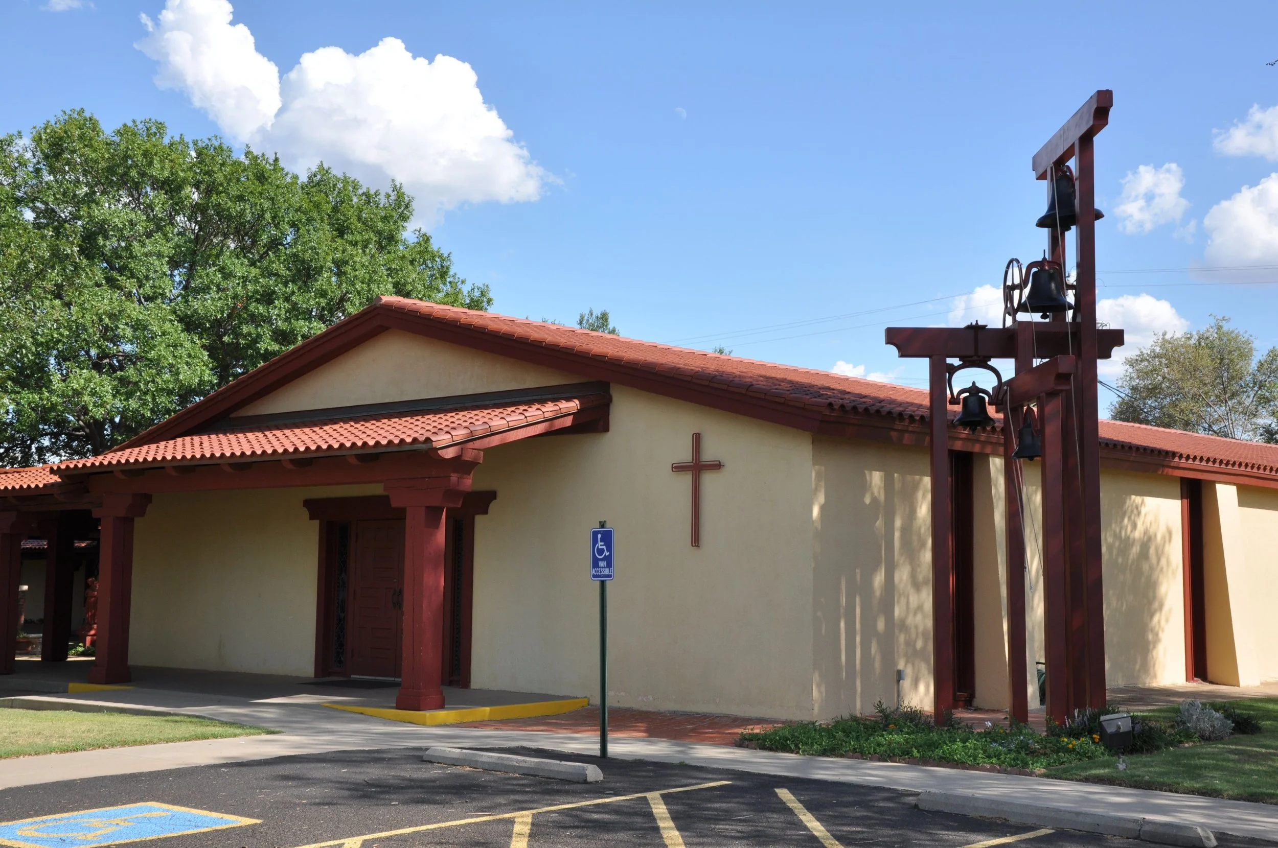 A church with a beige exterior and red roof, a cross on the wall, and a metal bell tower with three bells, situated in a parking lot with accessible parking space.