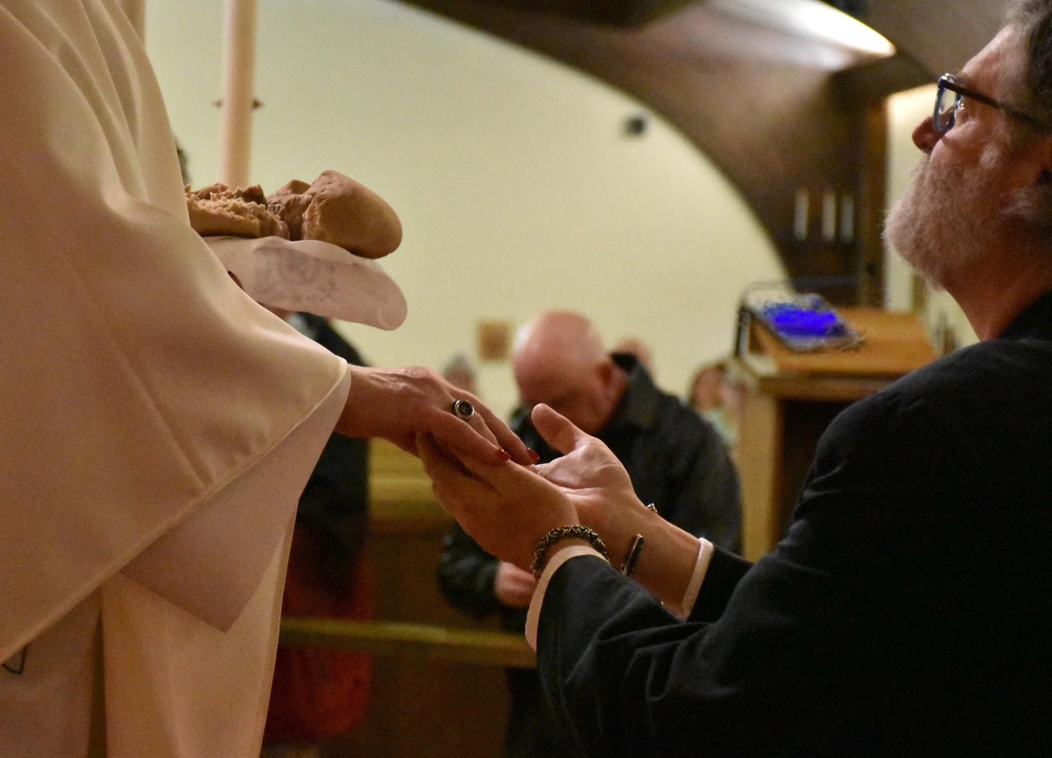 A man in glasses is holding hands with a woman wearing a beige dress during a wedding or formal event. The woman’s hand has a ring and bracelet, and the gesture appears tender and intimate. In the background, other guests are seated, and the setting is indoors, possibly a reception hall.