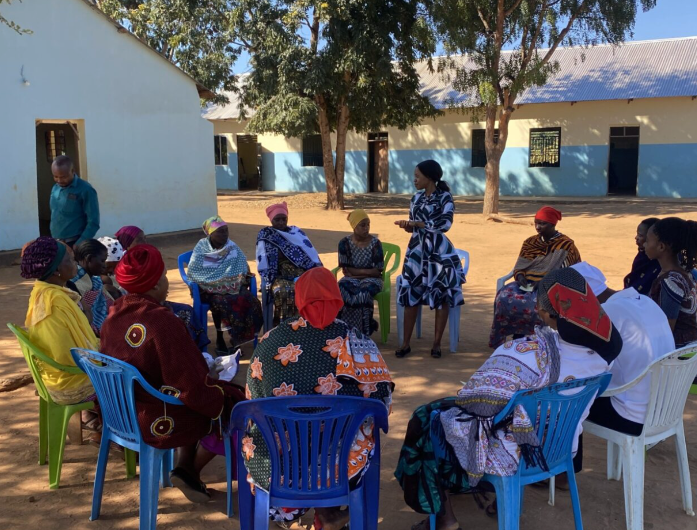 Women and children sitting on colorful plastic chairs outdoors in a circle, with one woman standing and speaking to the group, in a school courtyard with trees and buildings in the background.