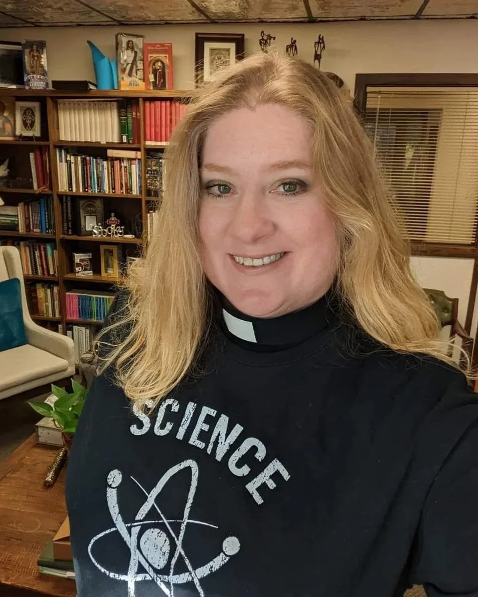 A woman with long blonde hair smiling, wearing a black t-shirt with a white atomic symbol and the word 'Science' on it, standing in a room with shelves filled with books and decorative items.