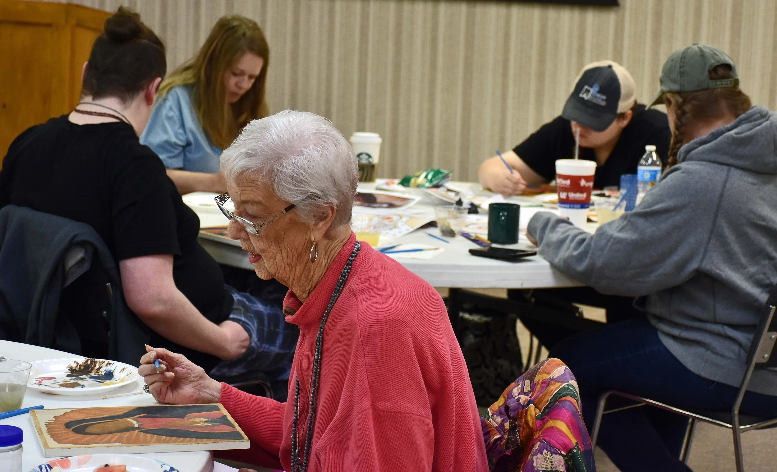 A group of people, including an elderly woman with white hair and glasses, sitting around a table working on art projects in a room with beige walls. The table has various art supplies and papers.