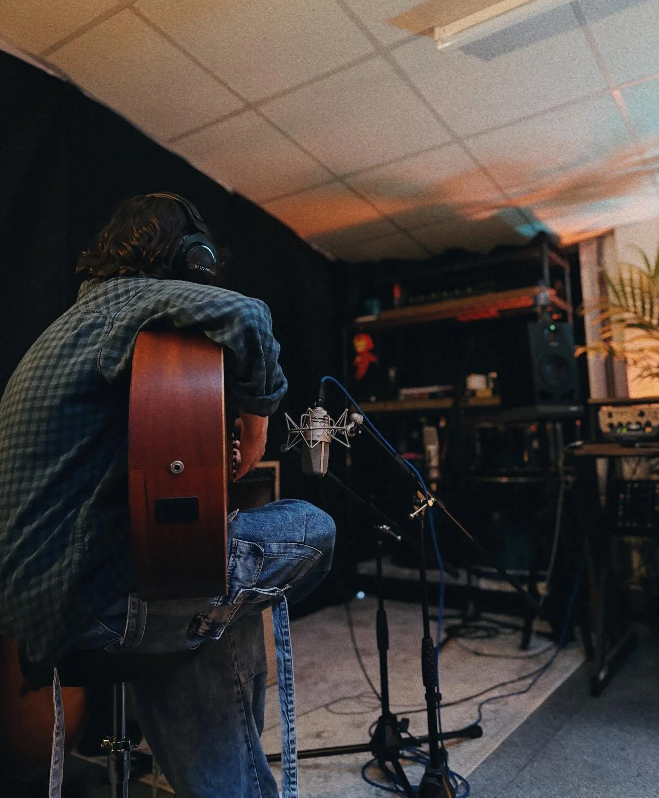 Person in a recording studio sitting on a stool with an acoustic guitar, wearing headphones in front of a microphone, with audio equipment and shelving in the background.