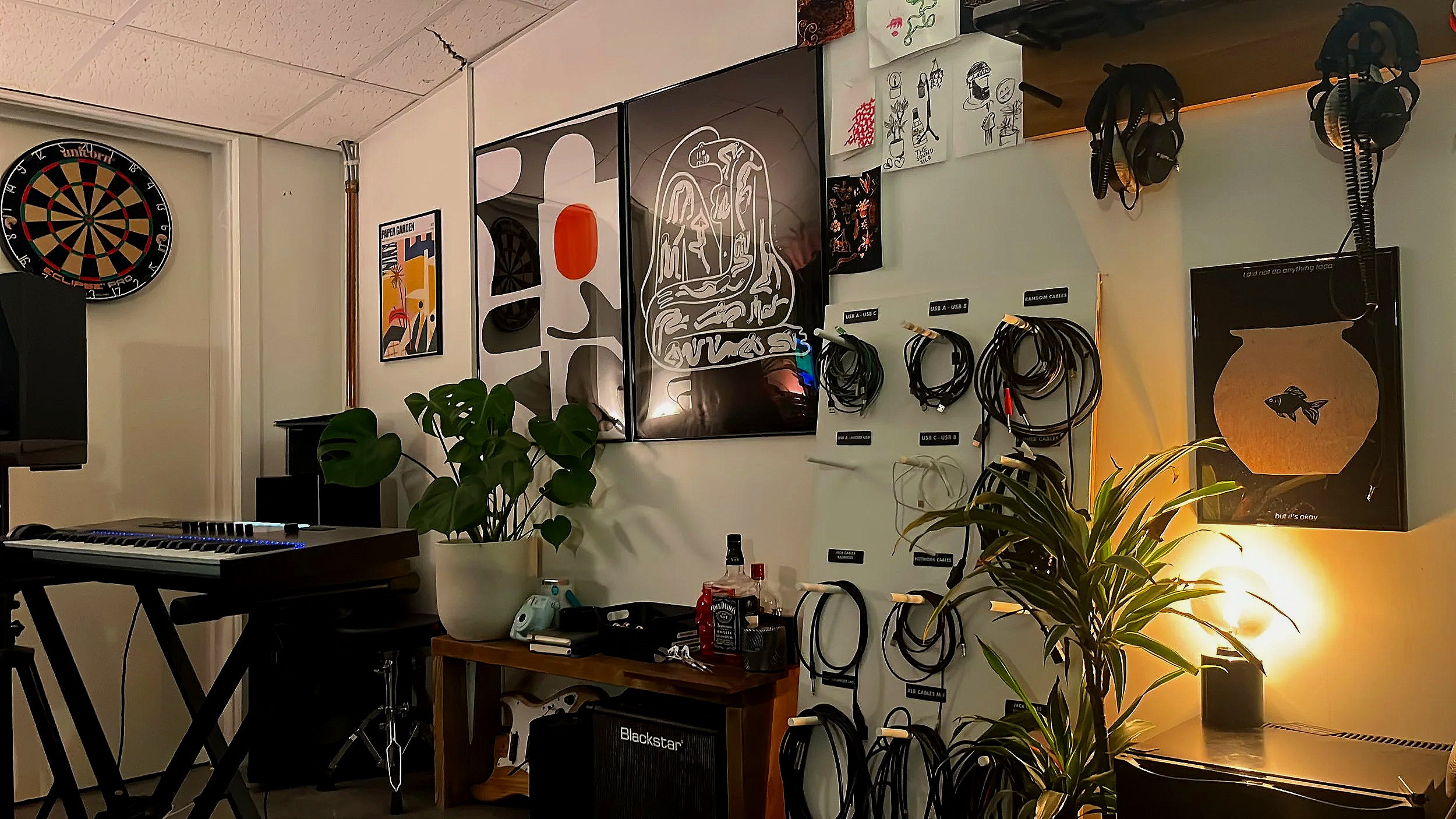 A cozy room with a dartboard on the wall, framed artwork, a digital keyboard on a stand, potted plants, a wooden table with bottles and a small speaker, and various cables hanging on the wall, with warm lighting and a lamp.
