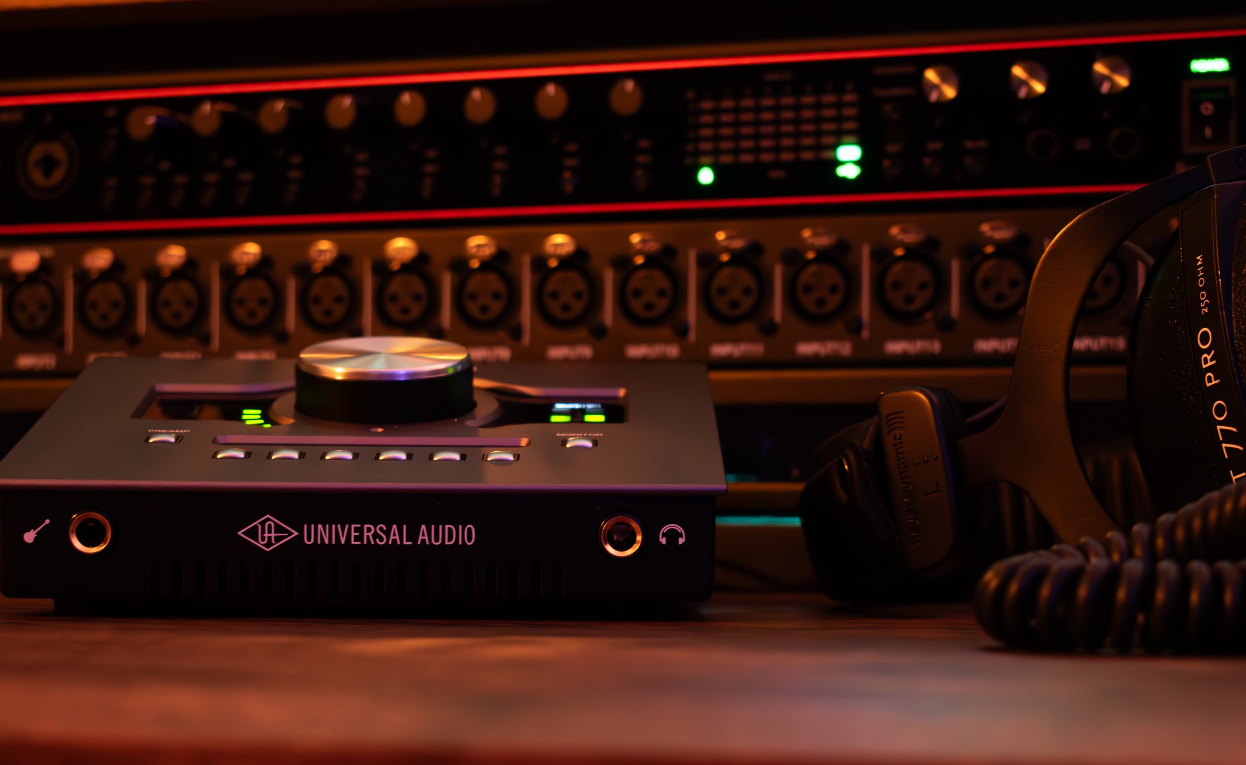 Audio equipment including a Universal Audio interface with a control knob, a pair of headphones, and a rack of audio gear in the background.